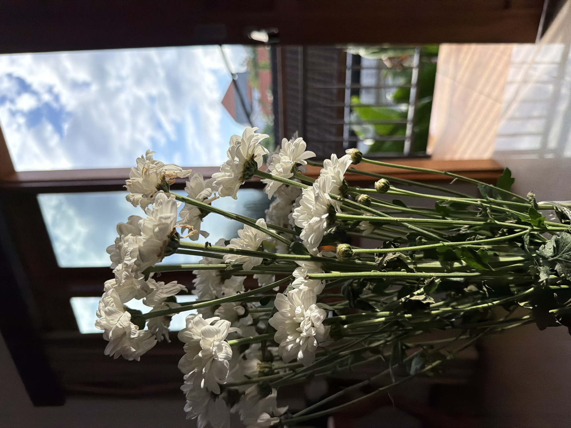 The image shows a bouquet of white chrysanthemums placed against a backdrop of an open window. Through the window, a cloudy sky is visible along with a partial view of buildings and greenery, indicative of an outdoor environment. The overall lighting suggests it is daytime. The image was taken in North Kuta, Indonesia.
