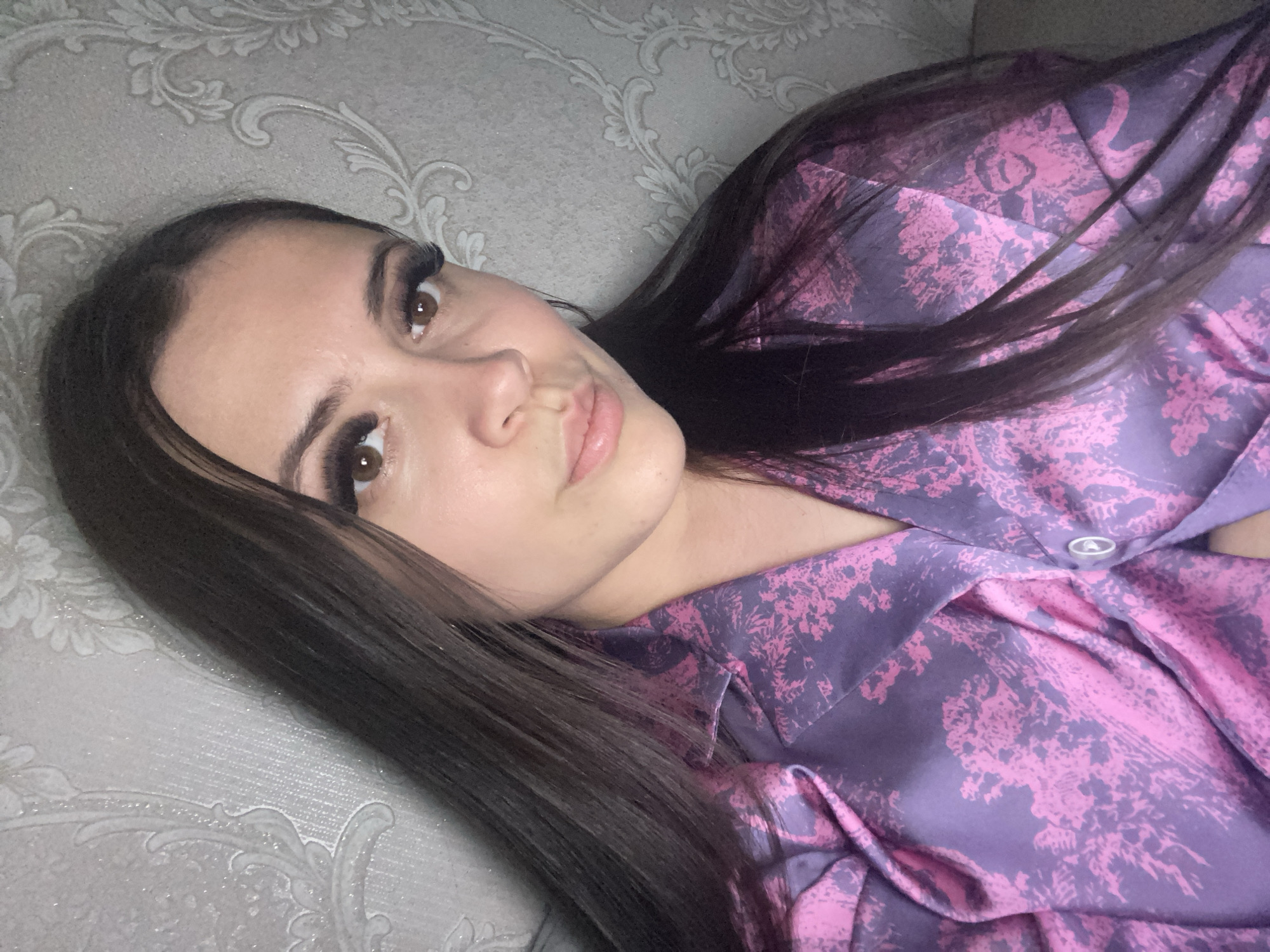 A young woman is depicted lying down, looking directly towards the camera in an indoor setting in Chaikovsky, Russia. She has long, dark brown hair, styled straight, which cascades over her shoulder and a patterned top. The top is made of a satiny fabric with a purple background and an abstract, vibrant pink pattern, resembling floral or botanical motifs. A single white button is visible near her collar. Her face features clear skin, defined dark eyebrows, prominent long eyelashes, and light brown eyes. Her lips are slightly parted and naturally colored, conveying a neutral and composed expression. The background is a light gray or beige wall covered in wallpaper with a subtle, shimmering damask-style pattern. The lighting is soft and even, suggesting an indoor environment during the day. No visible text is present.