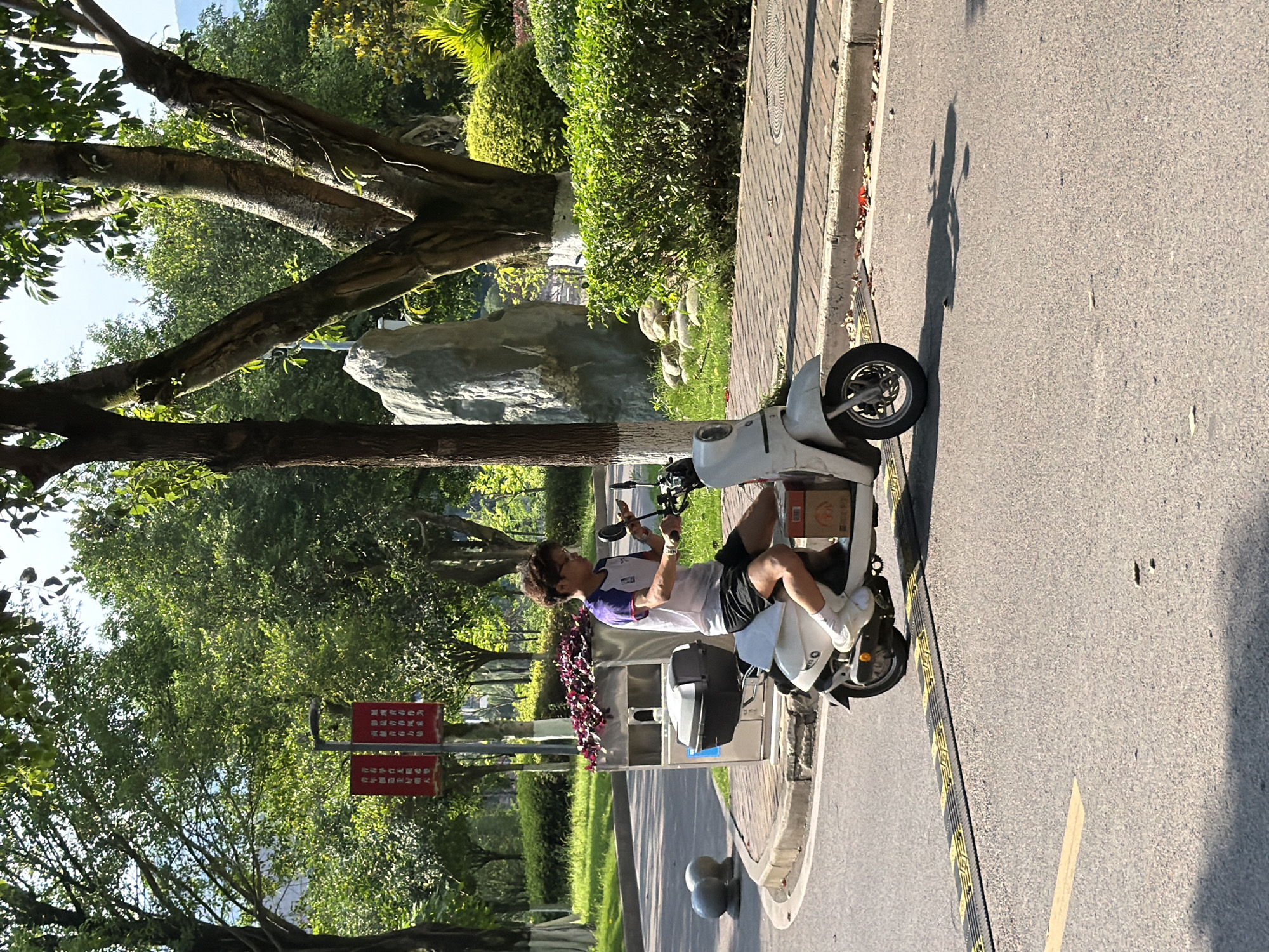 The image shows a person sitting on a white scooter on a paved road. The road is lined with trees and greenery. The location is likely Jiangjin, China, based on the signs and architecture visible. The person is holding a phone or device and appears to be interacting with it. The scooter has a box attached to the back. A raised walkway or curb separates the road from the greenery. The scene is outdoors during daylight hours.