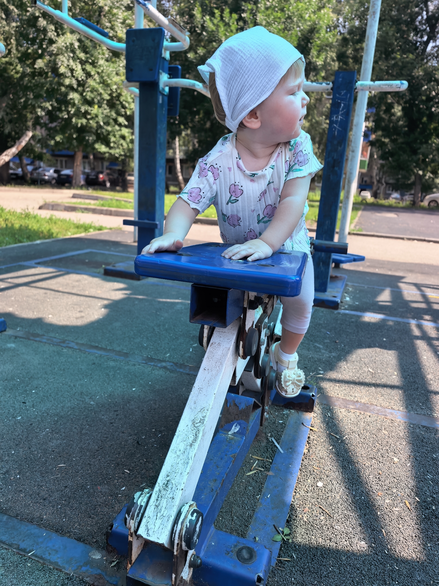 A young child wearing a white headscarf and a light-colored t-shirt with a floral pattern sits on a blue exercise machine in a park in Sterlitamak, Russia. The child is facing to the right, with their left hand resting on the machine's blue seat and their right foot on a pedal. The machine is blue and white, with visible metal components and rust. The ground is paved with dark, grainy material, and shadows from trees are cast across it. In the background, there are trees, buildings, and parked cars, indicating an urban outdoor setting. The lighting suggests it is daytime, possibly late morning or early afternoon, with bright sunlight.