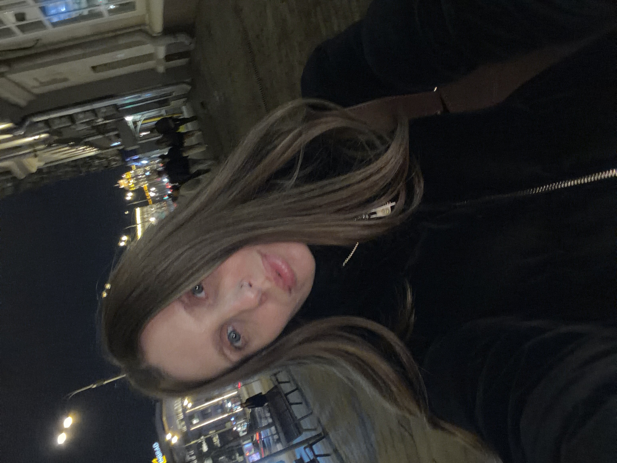 A young adult female with long, light brown hair and blue eyes, featuring a nose piercing on her right nostril, is depicted in a close-up selfie-style shot. She is wearing a black turtleneck top, a silver-colored chain with a rectangular pendant, and a black outer garment with a visible zipper pull on the left. A dark bracelet is visible on her right wrist.
The scene is set at night on an urban street in Moscow, Russia, indicated by a prominent partially visible yellow sign displaying "NGRAD" (likely part of "Leningrad" or a similar Russian name) on a building in the left background. The woman is on a paved sidewalk. To her left, a dark public bench is visible, with a solitary figure standing further down the street. In the background, streetlights illuminate the area, and several indistinct figures are seen walking along the sidewalk next to a multi-story building with light-colored facades and illuminated windows on the right. The sky is dark, and the overall environment is that of a bustling city at night.