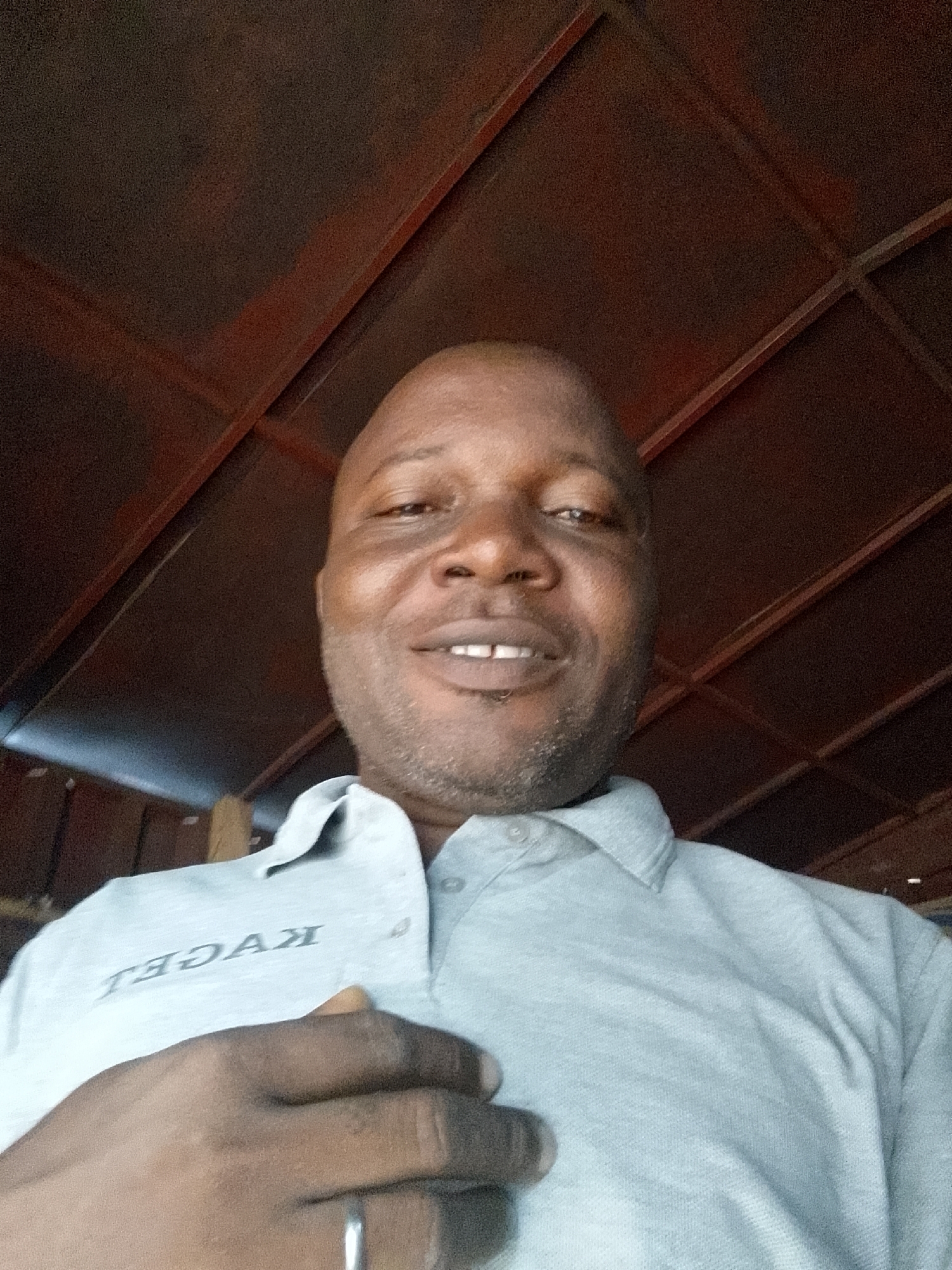 A smiling man is featured in a close-up, likely a selfie, looking directly at the camera. He is bald and has short stubble or a trimmed beard, revealing his upper teeth as he smiles. He is wearing a light grey polo shirt with dark grey capital letters printed on the left side of his chest. The text, which appears mirrored due to the likely selfie angle, reads "TEƆAƆEL" where the 'Ɔ' is a reversed capital 'C'. His left hand is placed over his chest, with a silver-colored ring visible on his left ring finger. The background is a dark reddish-brown ceiling or overhead structure, characterized by a grid-like pattern of beams or frames. The man's face is well-lit, contrasting with the somewhat dimmer background.