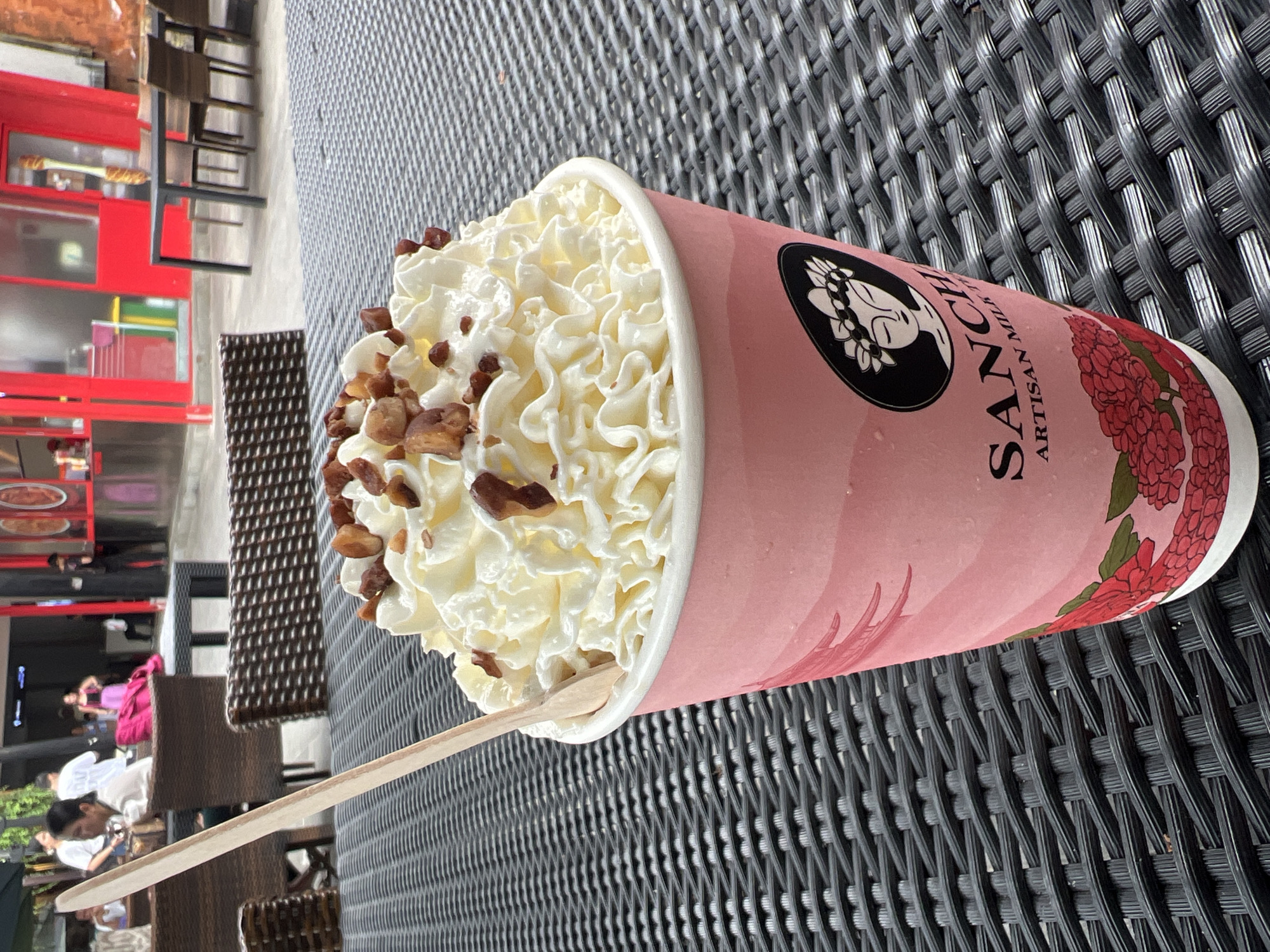 The image presents a large cup of what appears to be a milk-based drink with whipped cream and nuts on top, set on a black wicker table. A wooden spoon is inserted into the drink. The cup is pink, adorned with a floral design and the logo "SANCH ARTISAN MILK." In the background, there is a blurred scene showing people and a red building. Considering the branding and the location, the drink is likely being served at a cafe, possibly in the city of Kuta, Indonesia.