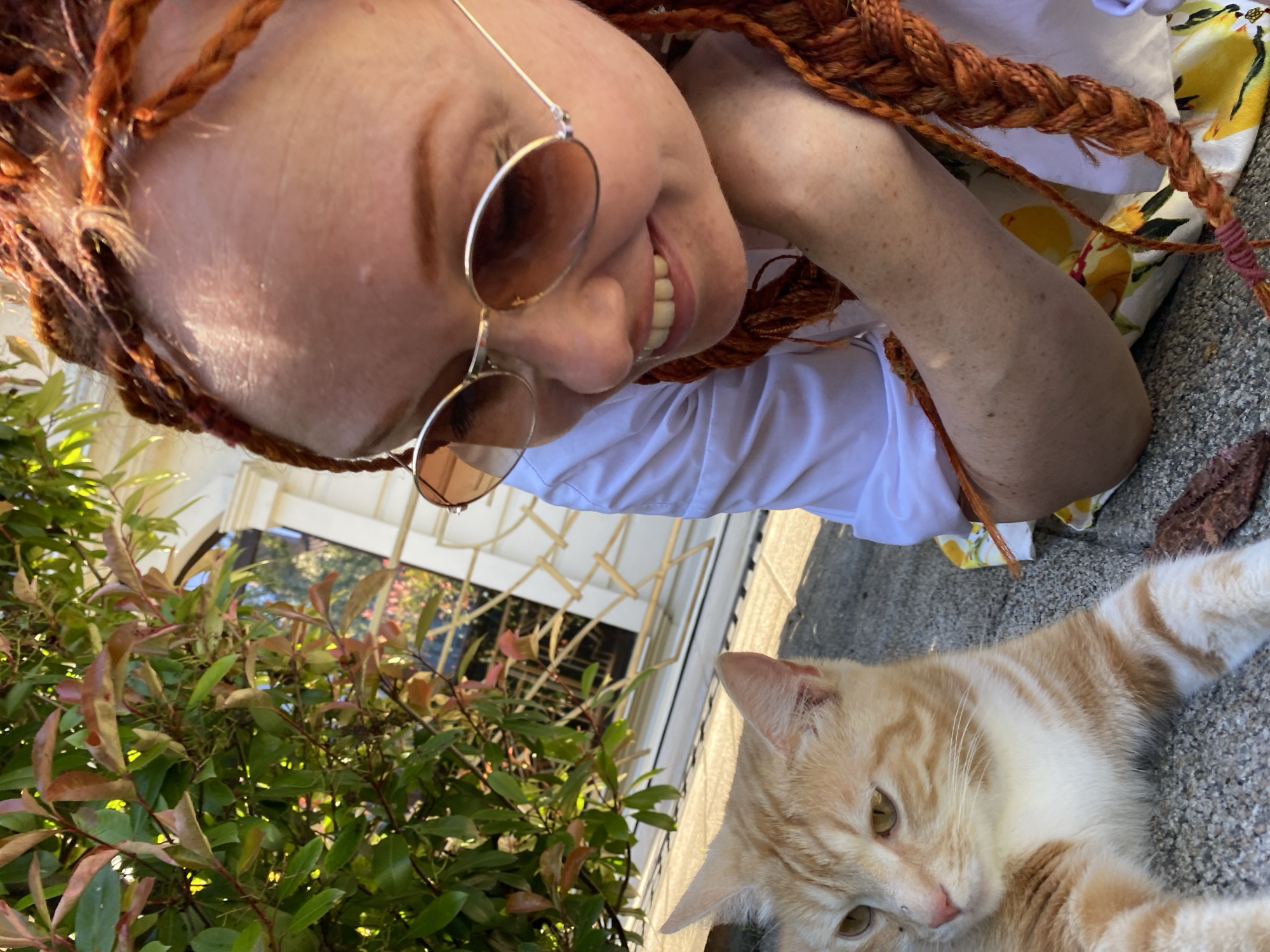 In this outdoor, close-up shot, a person with bright orange braided hair is tilted towards the camera, their face angled upwards. They are wearing round, rose-tinted sunglasses. Their mouth is open in a smile, revealing white teeth. They are wearing a white collared shirt, and part of a yellow and white patterned fabric is visible. To the right, an orange and white tabby cat is lying down on a sandy or pebbly surface, looking towards the camera with a relaxed expression. The background shows lush green foliage on the left and a white structure with lattice work on the right, suggesting an outdoor patio or garden setting. The lighting appears to be natural daylight, likely late morning or early afternoon given the brightness and shadows. The overall mood is casual and friendly. No visible text is present in the image.