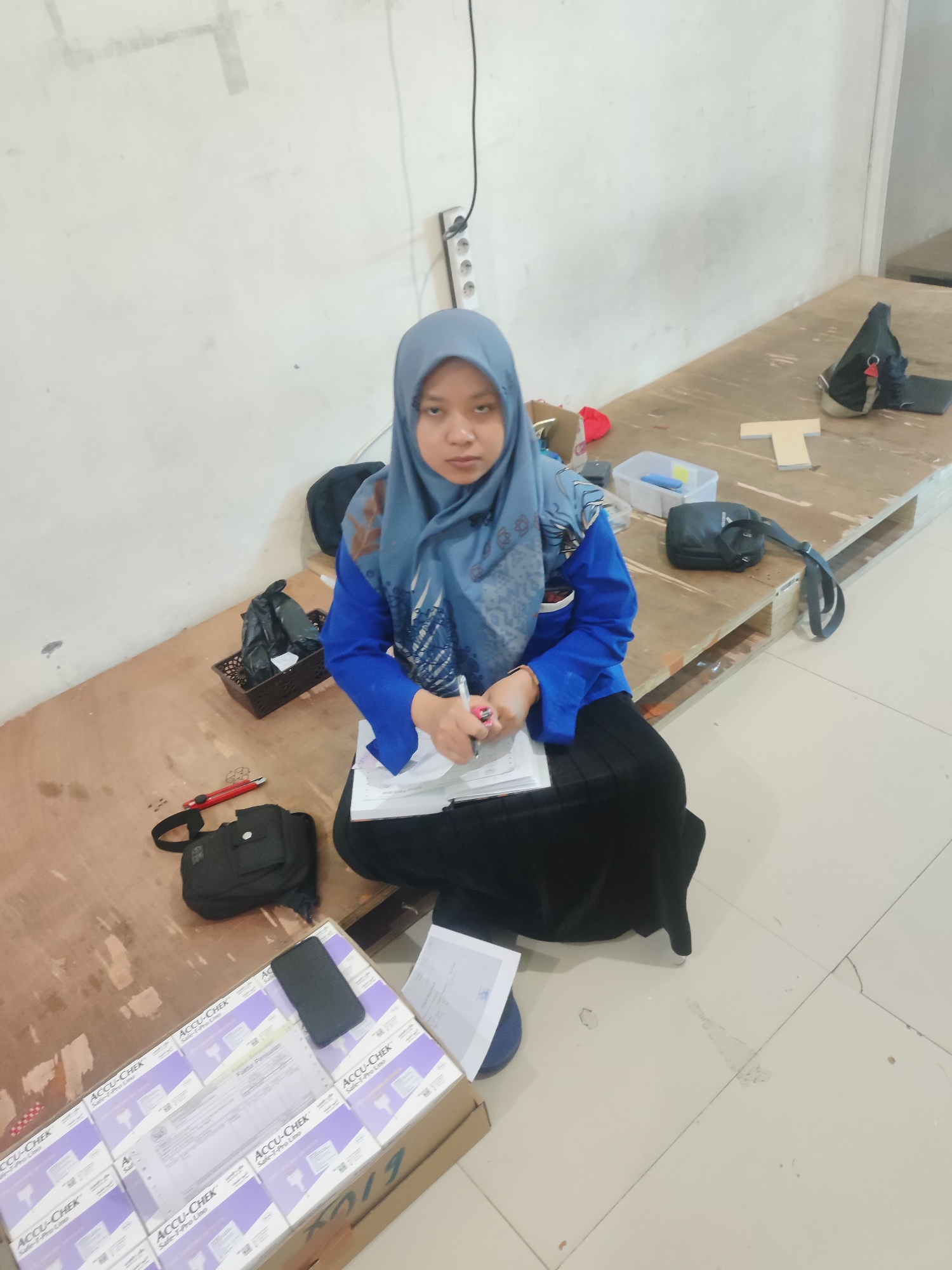 A young woman wearing a blue hijab and a blue shirt sits on a wooden pallet in a room with white walls and tiled floors. She is looking at the camera and writing on a stack of papers. In front of her is a black bag, a red utility knife, and a box filled with smaller boxes of "Accu-Chek Safe-T-Pro Uno" test strips. To her right, there is another black bag with a strap. The room appears to be a storage or office space. The overall scene suggests a work or study environment.