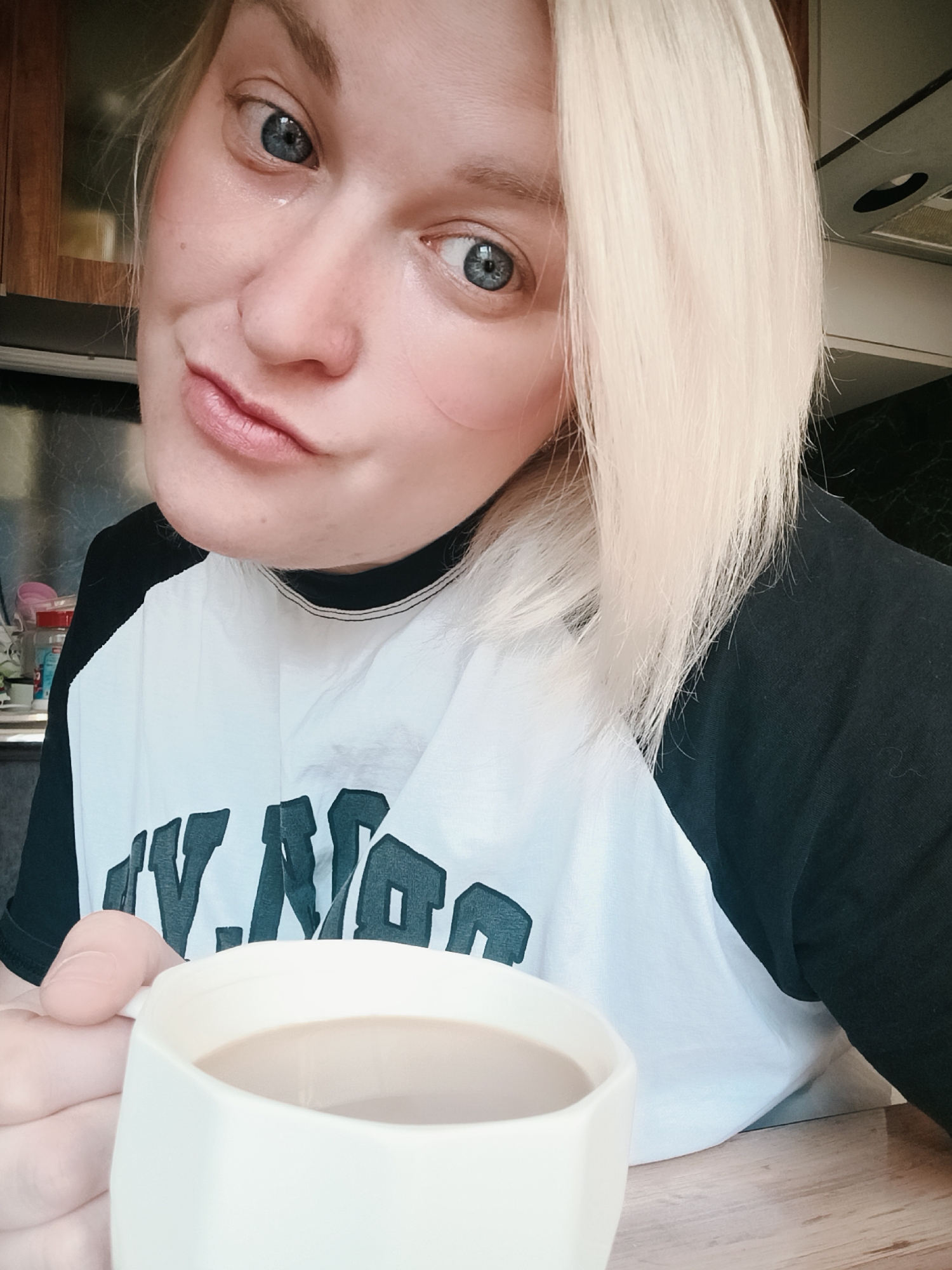 A woman with blonde hair and blue eyes is holding a white mug filled with a light brown liquid, likely coffee with milk or creamer. She is wearing a black and white t-shirt with text on it. The background shows a kitchen setting with wooden cabinets and a stainless steel backsplash. The location is likely in an interior space in the city of Chaikovsky, Russia.