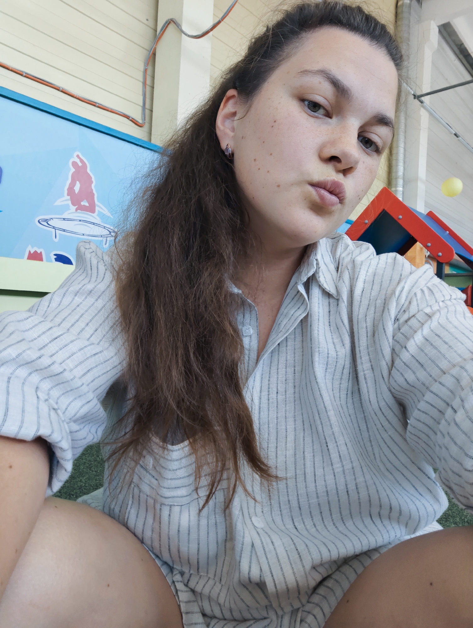 A woman with long, dark brown hair is looking at the camera and puckering her lips. She is wearing a light-colored, pinstriped shirt. The background appears to be an indoor play area with colorful structures, a blue graphic depicting a person on a trampoline, and some electrical wiring visible on a light-colored wall. The scene is likely a children's entertainment center in Chaikovsky, Russia.