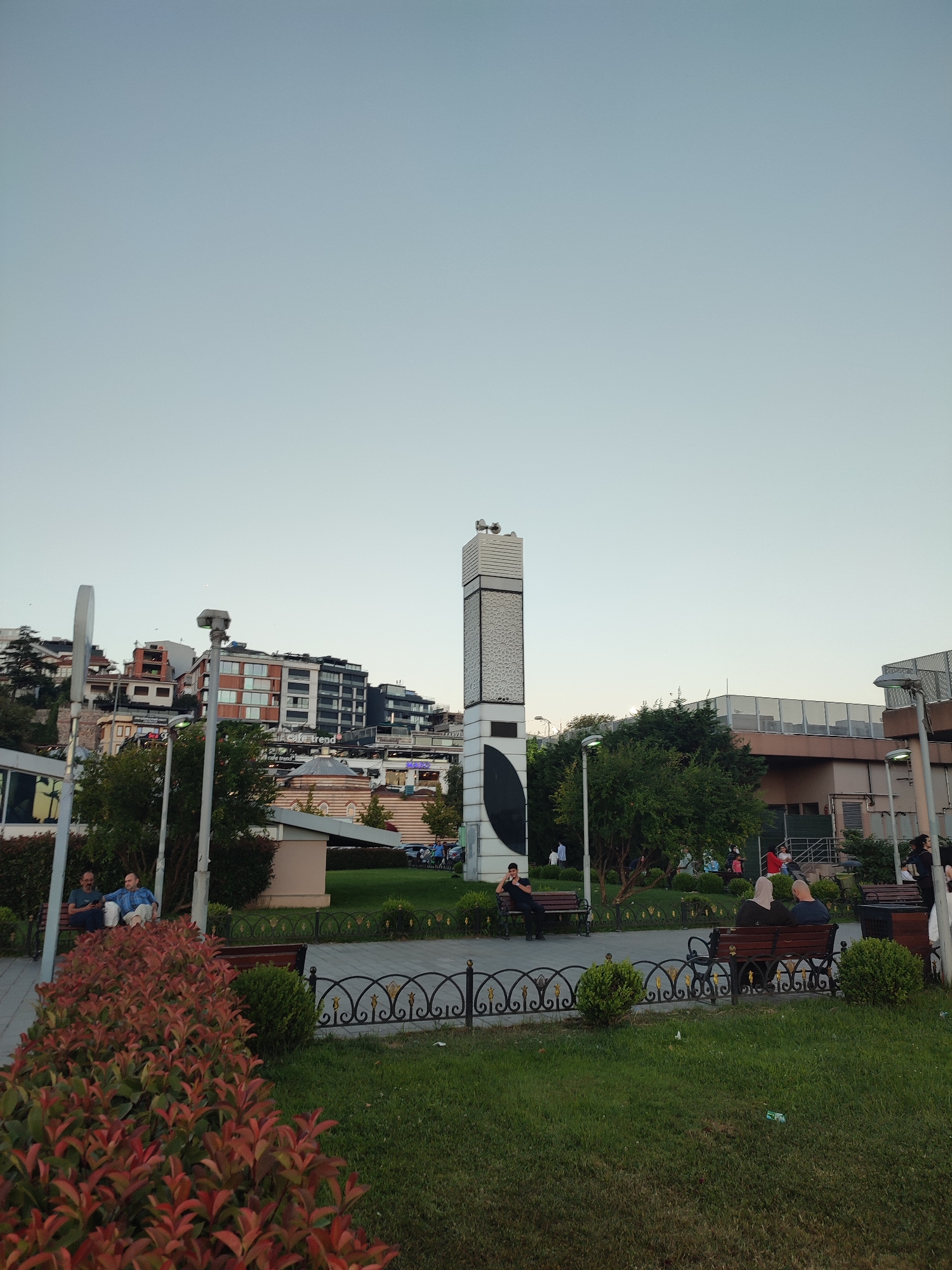 The image depicts a public square in Üsküdar, Turkey, with a prominent modern obelisk-like structure in the center. The sky is clear and a pale blue, suggesting it is either early morning or late afternoon. In the background, a cluster of buildings with varying architectural styles, including some modern apartments and older structures with domes, are visible on a hillside.
In the foreground, a well-maintained park area features lush green grass, manicured bushes, and flowerbeds with reddish-brown foliage. Several people are seated on park benches, some conversing, others looking out at the surroundings. A decorative black metal fence with gold accents separates the paved walkway from the grassy area. Lampposts are positioned throughout the park.
To the left, a pathway leads towards the buildings, and some vehicles are parked in the distance. The overall atmosphere appears peaceful and relaxed, with people enjoying the outdoor space. The obelisk, with its geometric patterns and a large dark circular emblem near its base, is the focal point of the scene.