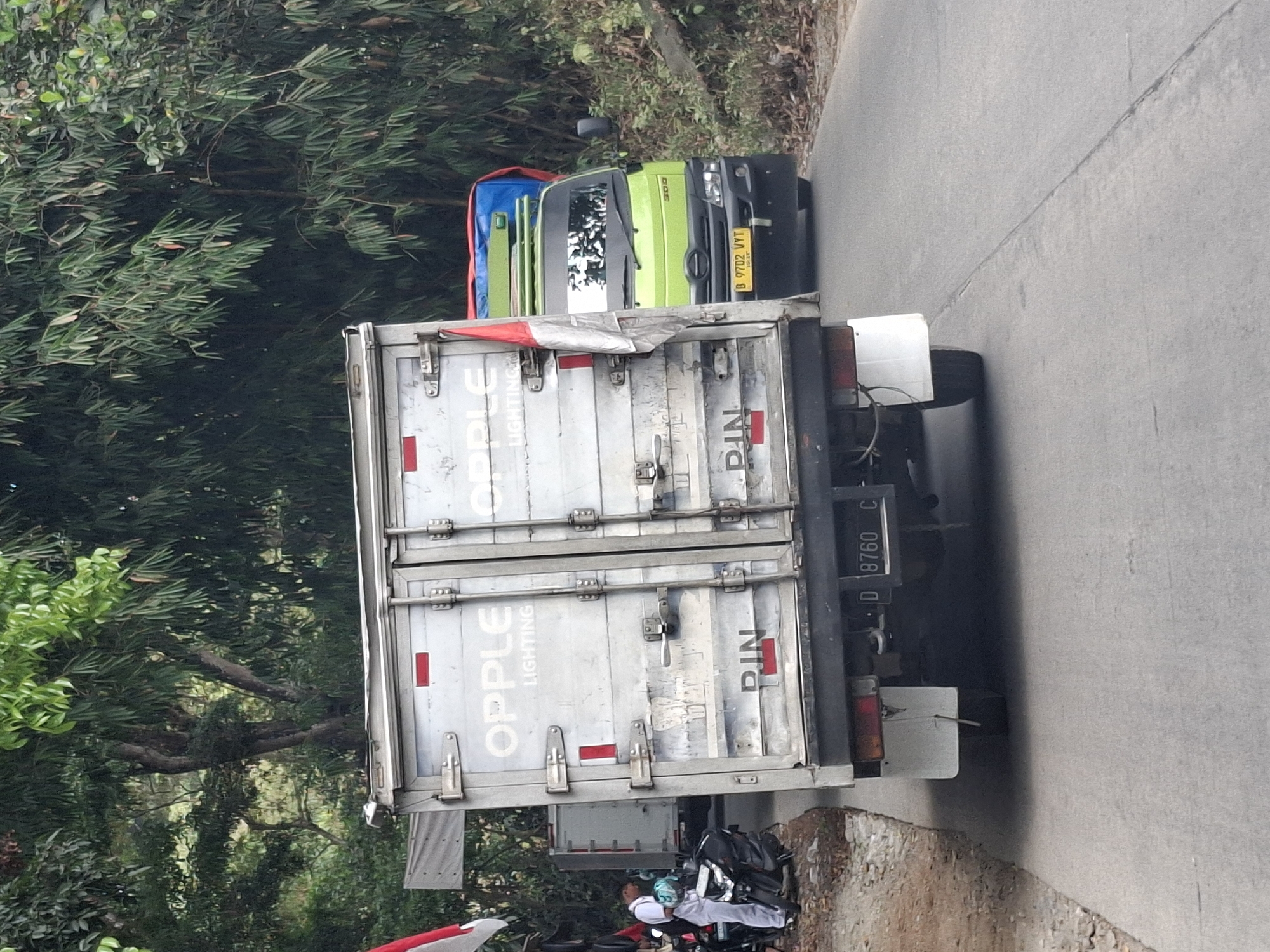 A two-axle truck, identified by the license plate D 8760 C, is parked on a paved road in Cibadak, Indonesia. The truck is silver with "OPPLE LIGHTING" printed on its side. A red and white Indonesian flag is attached to the truck. To the right of this truck, a green truck with license plate B 9702 VYT is visible. In the background, there are trees and foliage. A person in a white uniform is standing next to a motorcycle on the left side of the road. The scene appears to be a roadside setting.