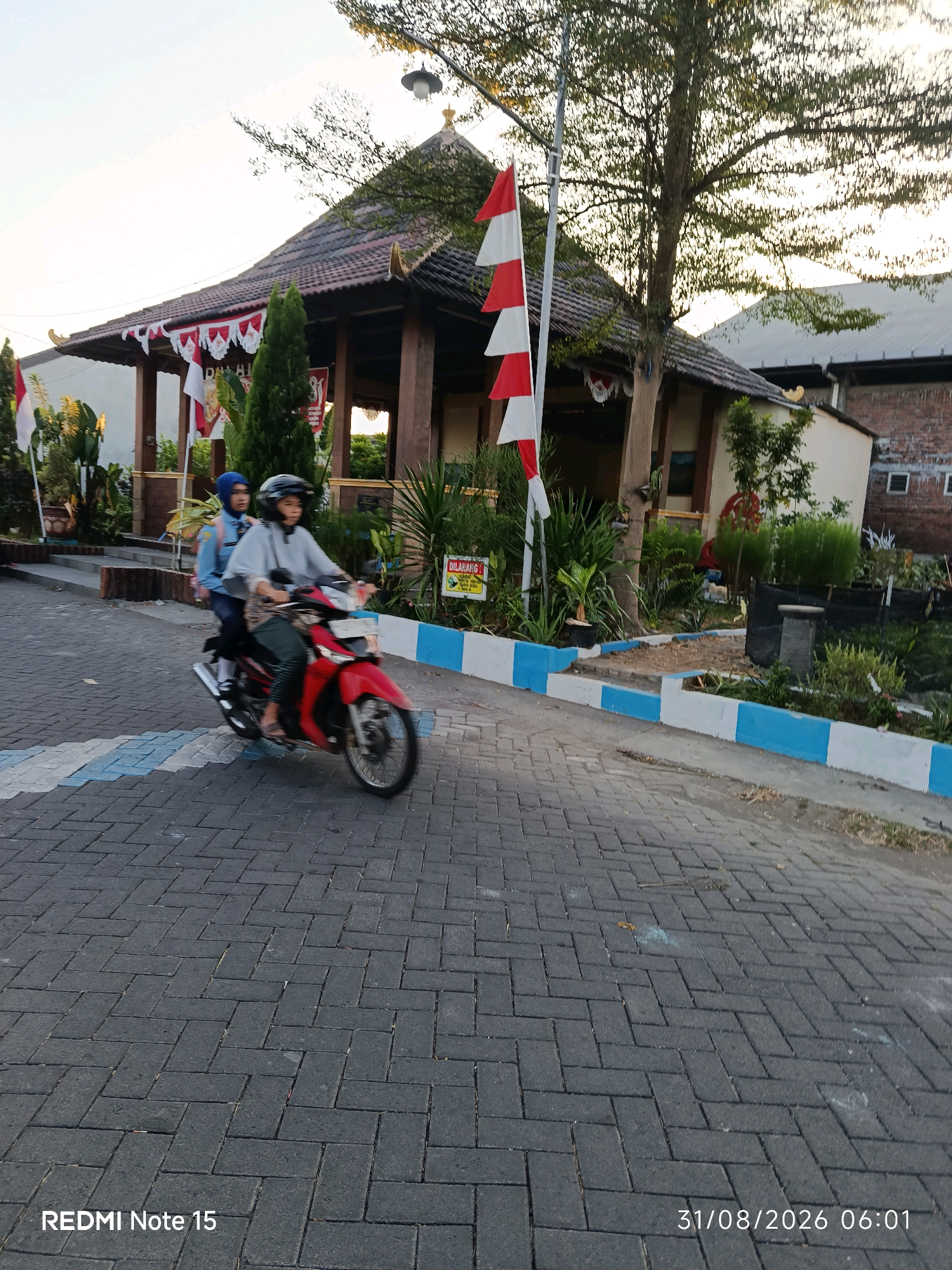 Two individuals, one driving a red motorcycle and the other as a passenger, are depicted in motion on a brick-paved road. The driver is wearing a helmet and a patterned skirt, while the passenger is wearing a blue headscarf and a blue shirt. They are positioned in the foreground, with the motorcycle facing to the right. In the background, a traditional Indonesian-style building with a tiered roof is visible. Red and white flags are displayed along the building's eaves and on a flagpole. The environment appears to be an outdoor setting, possibly a public area or a school campus, with trees and greenery surrounding the building. The sky is overcast, suggesting it may be early morning or late afternoon. The timestamp "31/08/2026 06:01" is present in the lower right corner of the image, indicating the date and time the photograph was taken. The location is identified as Keboansikep, Indonesia.