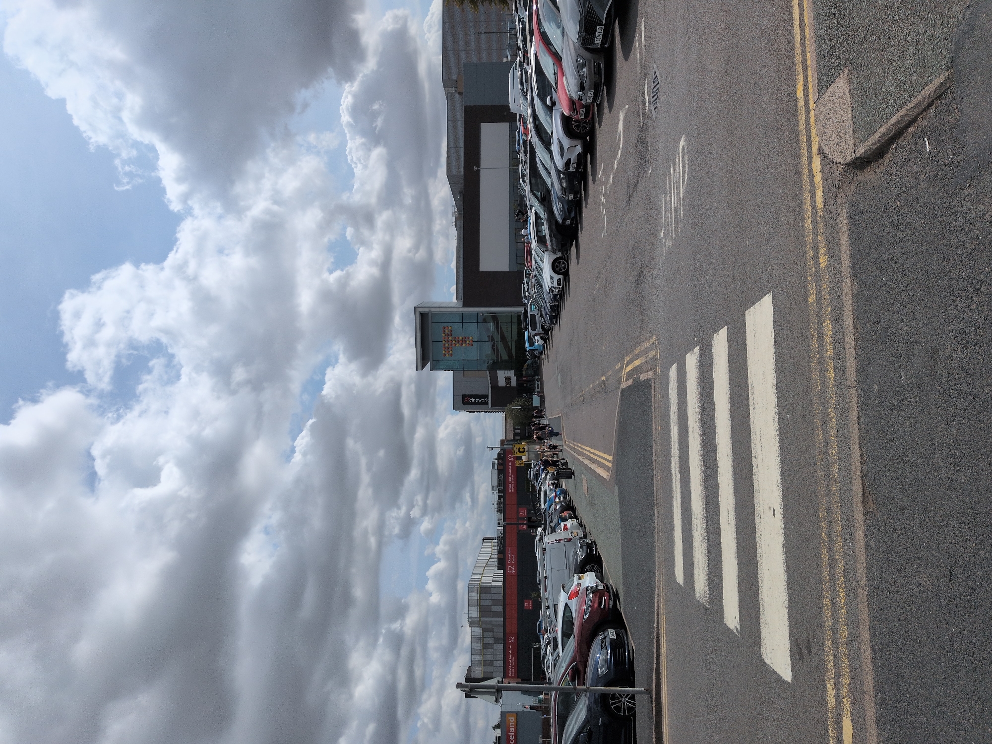 A vertical, eye-level, medium shot captures a wide, paved parking lot with a pedestrian crossing prominently in the foreground. The parking lot stretches towards a row of buildings under a cloudy sky. The sky is dominated by large, grey, cumulus clouds with patches of blue sky visible. A white pedestrian crossing with yellow double lines on either side marks the immediate foreground. Beyond the crossing, rows of parked cars, predominantly sedans and SUVs, fill the parking lot, with vehicles in various colors including black, grey, white, red, and blue. A few people, dressed in casual clothing, are walking in a group near the far end of the parking lot, heading towards the buildings. The buildings in the background are modern, with glass facades and some featuring illuminated signage. One prominent building on the left has a sign that reads "Iceland". Another building on the right has a large "T" logo with an orange glow. The scene appears to be a commercial area, likely a retail park or shopping center, in Telford, United Kingdom. The overall impression is a bright, slightly overcast day in an urban commercial setting.