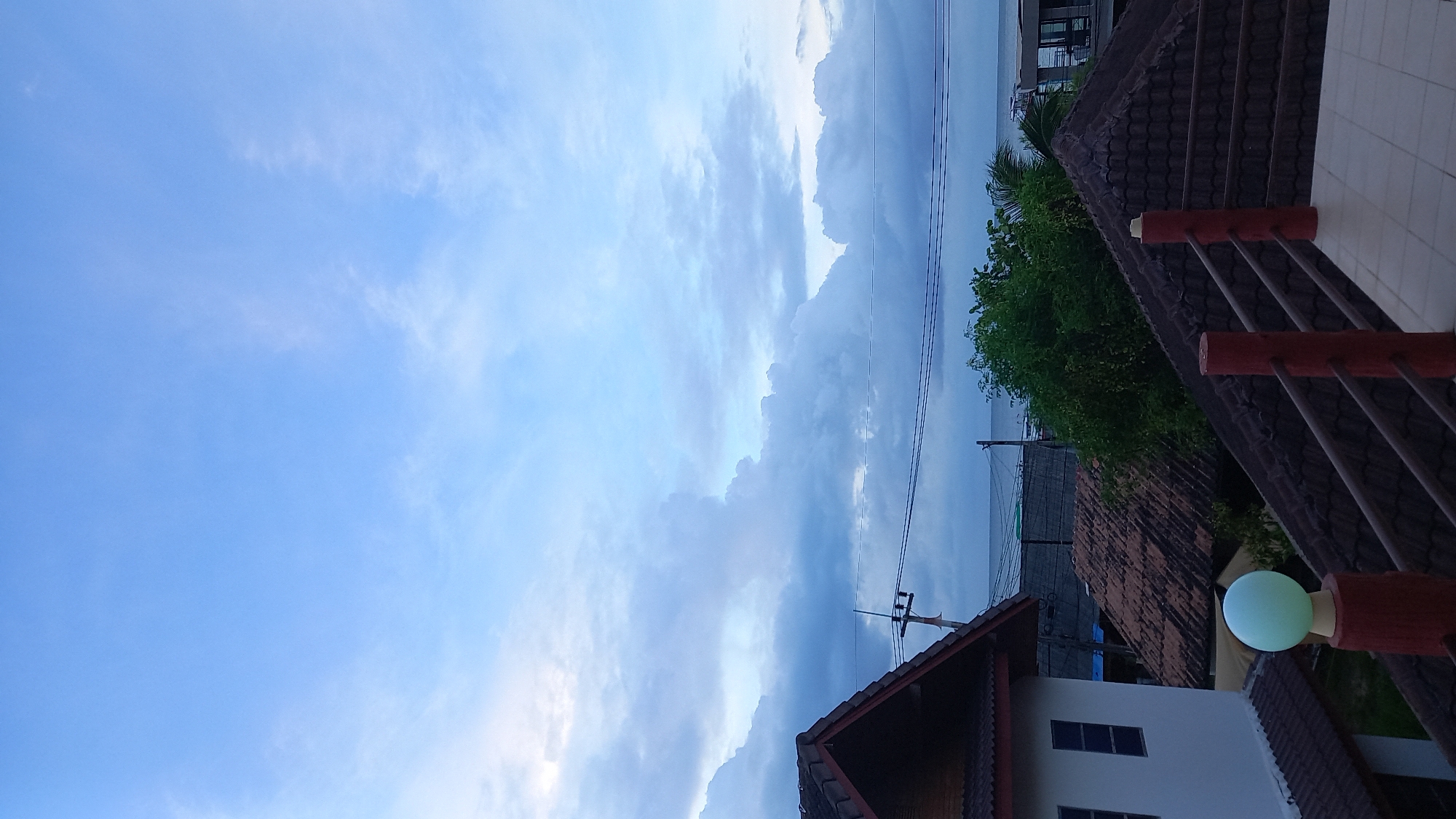 The image depicts a view from a rooftop in the city of Ko Pha-ngan District, Thailand, overlooking a residential area near the sea. The sky is filled with a mixture of blue and cloudy formations, suggesting it is either early morning or late evening. In the foreground, a white house with a dark-tiled roof is partially visible on the left. Adjacent to it are other buildings with terracotta-colored roofs. A light fixture with a round, pale green globe sits on a red post in the lower right quadrant of the image. A railing, also with red posts and metal bars, lines the edge of the rooftop. Beyond the buildings, a lush green tree stands out, and further in the distance, the calm sea is visible, with a few boats or structures on the horizon. Power lines and a utility pole are also present, crossing the view of the sea. There are no people visible in the scene. The overall impression is one of a quiet coastal town.