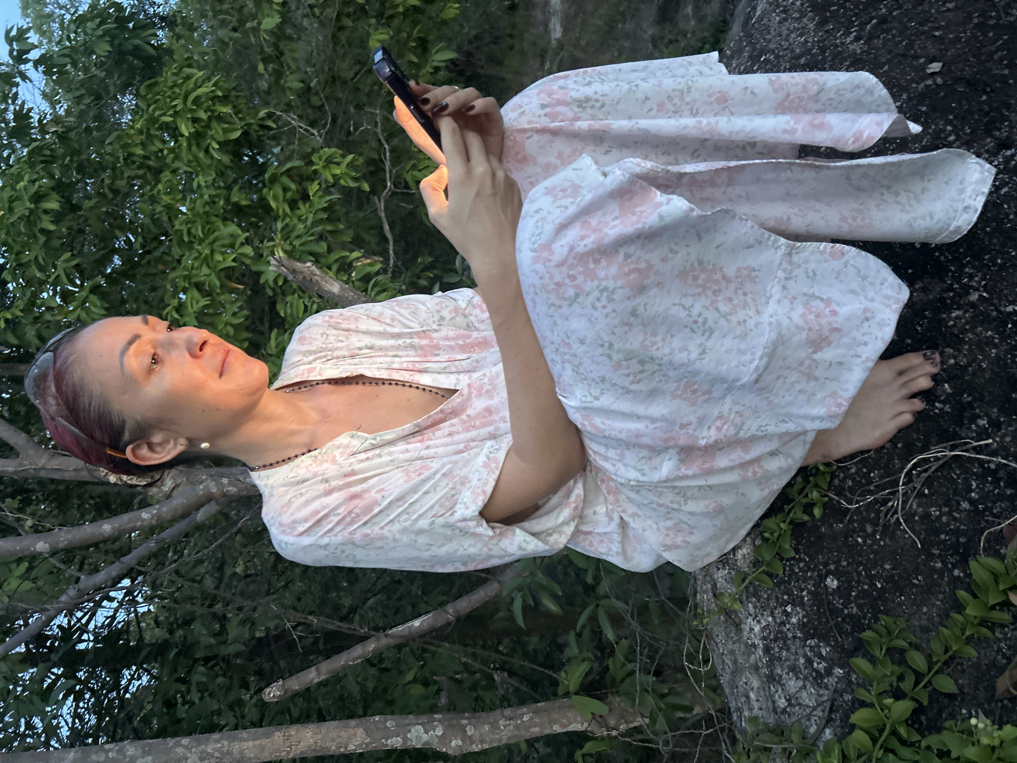A woman is sitting on a rock in an outdoor environment in Baan Hin Kong, Thailand. She is wearing a light-colored, floral-patterned, long-sleeved dress or gown. She is holding a mobile phone and looking at the screen. The lighting on her face suggests the phone's screen is illuminated. Part of the scene includes trees and foliage surrounding the rock where she is sitting. The rock appears to be covered in vegetation.