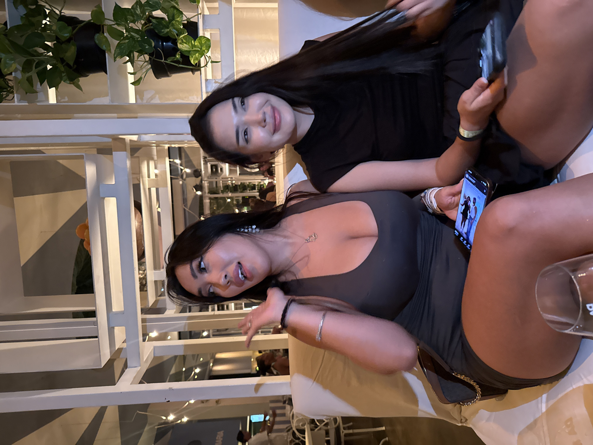 Two young women are seated on a cream-colored sofa within an indoor setting. The woman on the left is wearing a dark grey, fitted dress and is gesturing with her left hand while looking to her left. She has long, dark hair, visible cleavage, and is wearing jewelry including earrings and bracelets. A dark purse with a gold chain rests on her lap. The woman on the right is wearing a black, sleeveless top and a black skirt. She has long, dark hair and is smiling while looking to her right. Both women are holding smartphones. The background features white shelving units with potted plants and decorative elements, suggesting a restaurant or lounge environment in North Kuta, Indonesia.