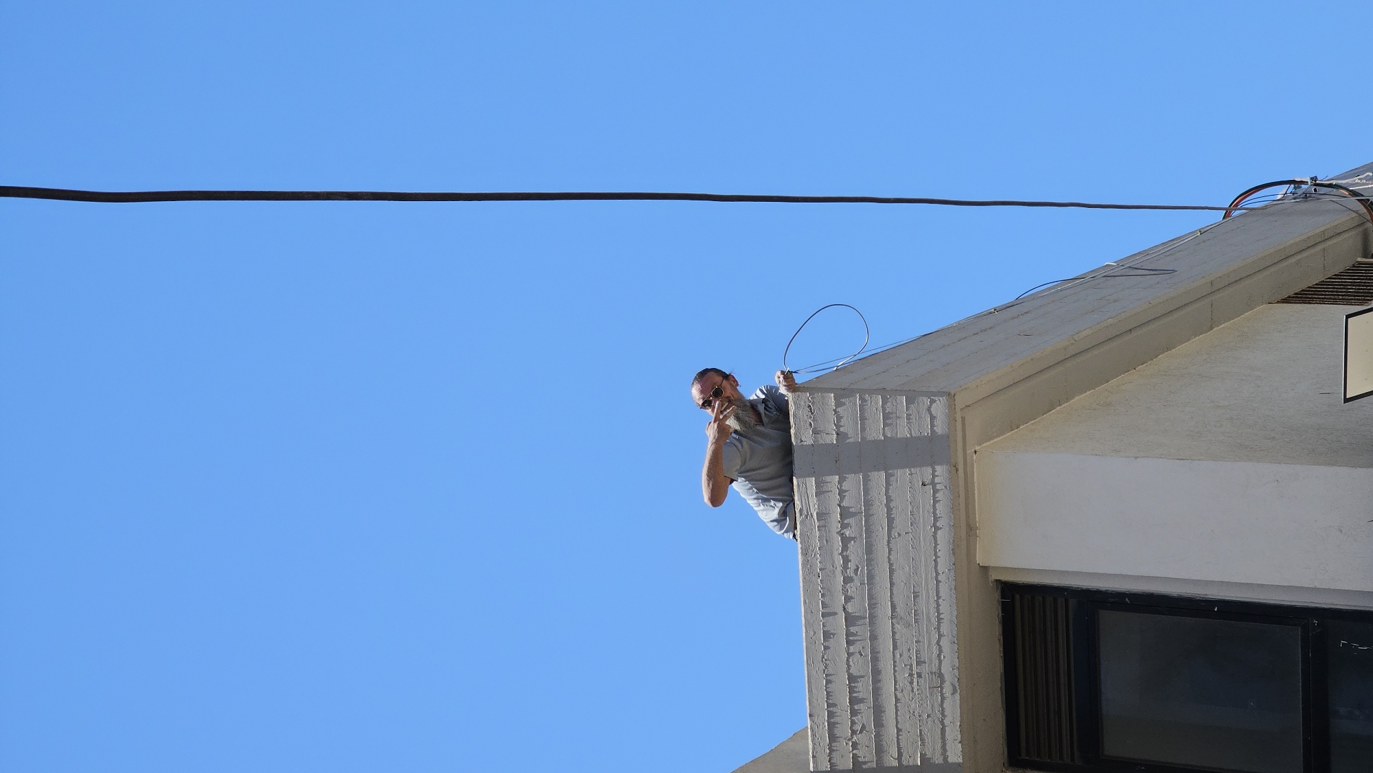 A man with a beard and sunglasses is leaning over the edge of a concrete rooftop against a clear blue sky. He is wearing a light gray t-shirt and is holding a coiled black cable in his right hand, with his left hand extended towards it. A long, thin black cable runs vertically from the upper right of the frame down along the side of a white building. The building has a textured concrete edge and a dark-framed window with horizontal blinds. The image was taken in Holon, Israel.