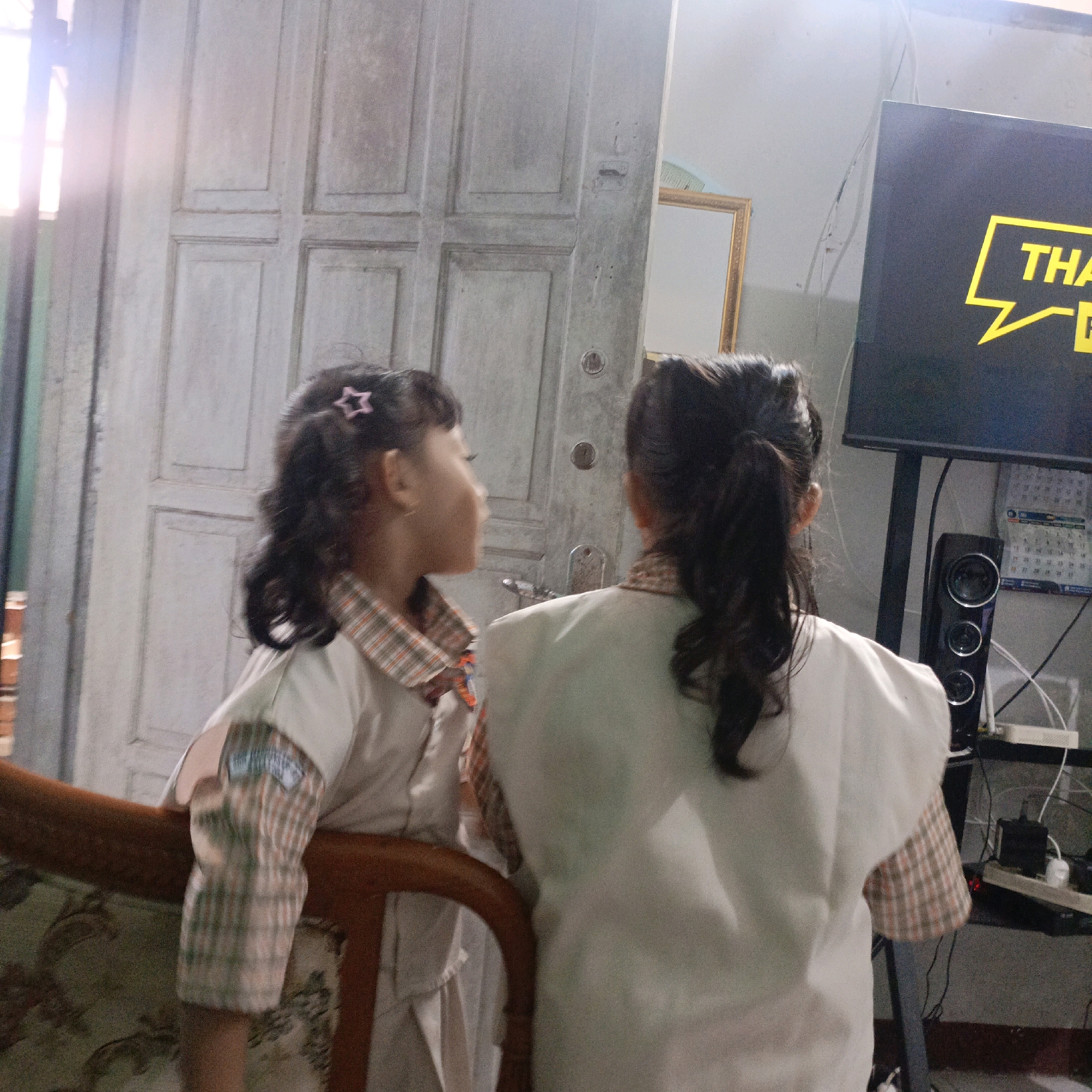 In a room in Salatiga, Indonesia, two young girls are seen from behind and the side. The girl on the left has dark, curly hair with a pink star-shaped clip and is wearing a light-colored uniform with plaid sleeves. She is looking to her right. The girl on the right has dark hair tied back and is wearing a similar uniform. They are standing near a light-colored, paneled door. In the background, a computer monitor displays a yellow graphic with the word "THA" and a speech bubble. A black speaker sits on a shelf with a calendar and other electronic devices. The room appears to be dimly lit, with light coming from the left, possibly a window.