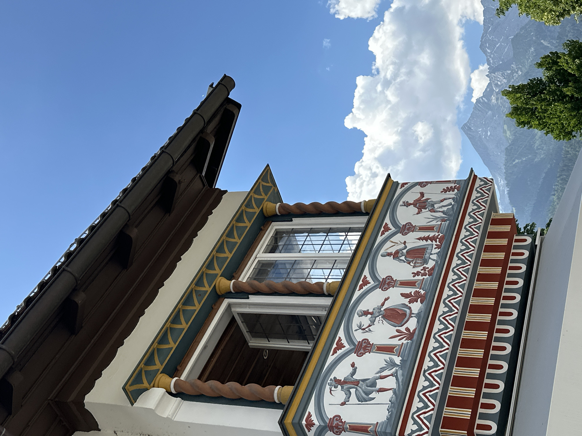 The image captures a close-up of a building's facade in Garmisch-Partenkirchen, Germany. The focus is on a decorated balcony with colorful paintings depicting figures in traditional attire, possibly from Bavarian folklore. Twisted wooden columns support the roof. The architecture is ornate, with a scalloped roof edge and a detailed frieze. Beyond the building, a majestic mountain range under a bright blue sky with scattered white clouds provides a scenic backdrop. The scene suggests a picturesque and historic setting, characteristic of the Bavarian Alps region.