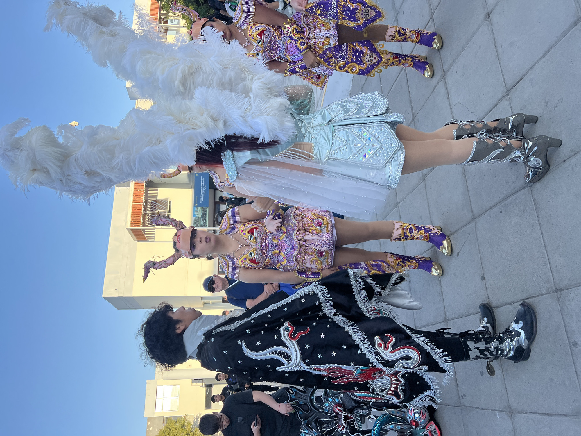 In Arica, Chile, four individuals in elaborate traditional costumes are gathered on a paved public walkway. The central figures, two women and one man, are adorned in vibrant, sequined garments, ornate headwear, and tall boots, suggesting a cultural performance or festival. The woman on the far left wears a striking black cloak embellished with a silver snake design and white fringe. She stands facing a woman in a purple and gold costume topped with a horned headdress. Another woman, to the right, is dressed in a pale blue, shimmering outfit with large white feathered wings extending from her head and shoulders. Behind them, other individuals in casual attire and a building with architectural details are visible. The scene is set against a clear blue sky, with sunlight illuminating the festive attire.