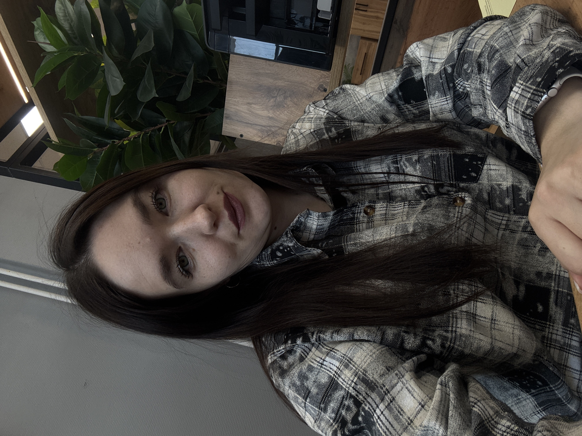 A young woman with long, dark brown hair is captured in a close-up photograph, seemingly at a cafe. She is wearing a plaid shirt with a color scheme of white, gray, and black. Behind her is a lush green plant. In the background, elements like furniture and structural beams suggest an indoor setting. The location may be in the city of Chaikovsky, Russia.