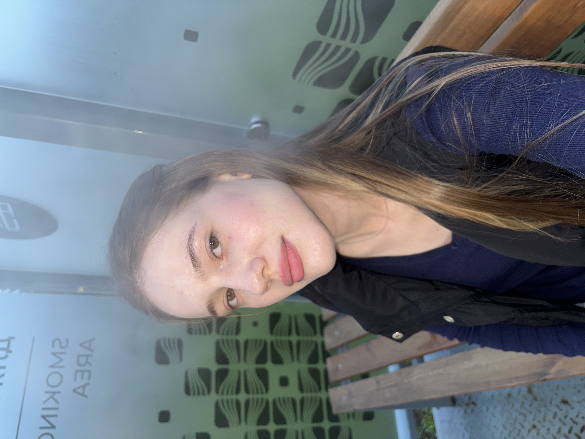A young Caucasian woman with light skin, light brown hair, and brown eyes is captured in a selfie taken outdoors. She is wearing a navy blue long-sleeved shirt and a black vest over it. Her lips are painted with a pink lipstick, and she has a natural makeup look with rosy cheeks. The background consists of a green glass wall with a repeating pattern of wavy lines enclosed in squares, and a wooden bench. Above the woman's head, a sign is partially visible, with text that appears to read "SMOKING AREA" in mirrored, reverse lettering. The lighting suggests it is daytime, possibly overcast or in the shade, with a slight haze or mist present. The woman is seated on a wooden bench. The location is identified as the city of Arfa, Russia.