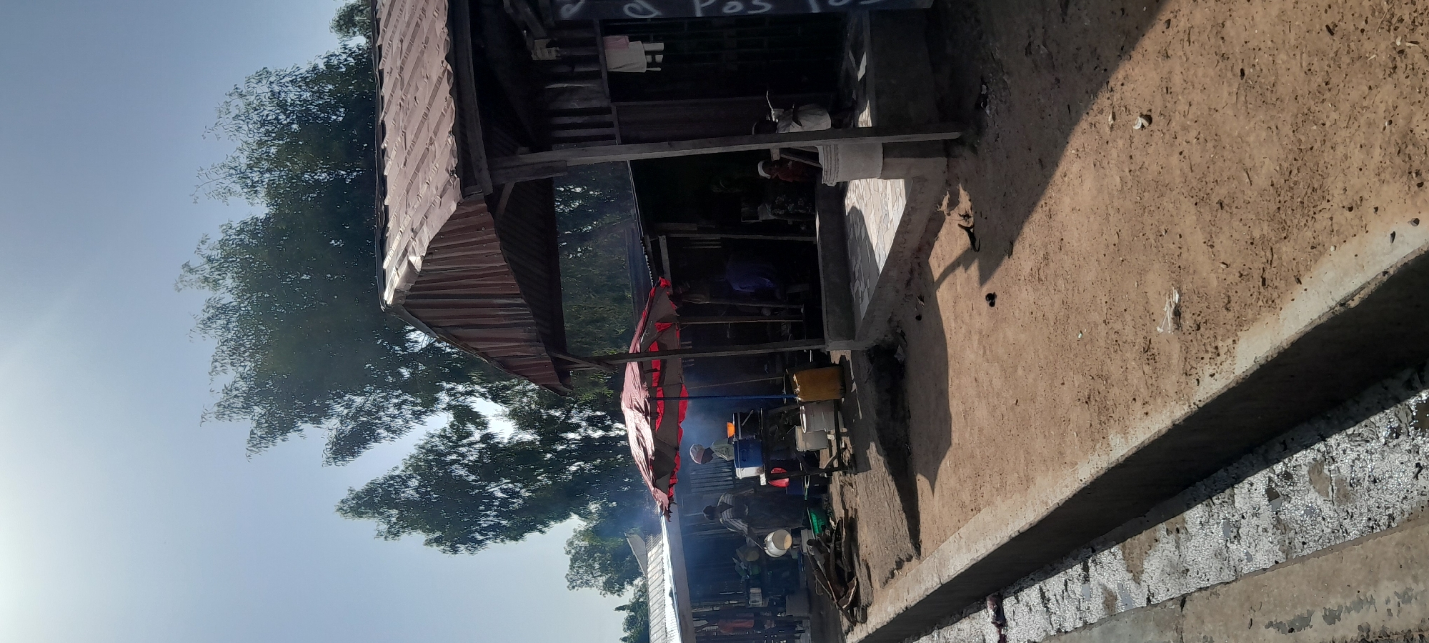 An outdoor market scene in Jalingo, Nigeria, shows several makeshift stalls under a bright, clear sky. The foreground features a dirt path next to a concrete drainage ditch. In the middle ground, a cluster of stalls are visible, some with corrugated metal roofs. Underneath a red umbrella, two people appear to be cooking or preparing food, with smoke rising. Other individuals are seen standing or seated near the stalls. The background consists of trees and a clear sky. The overall impression is a busy, functional marketplace.