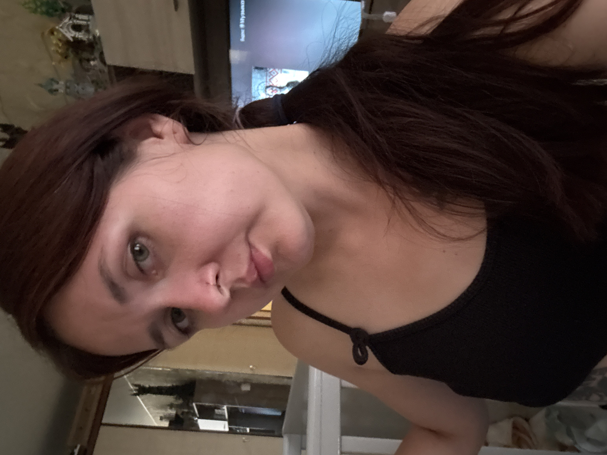 A young woman with brown hair pulled back in a ponytail and green eyes is looking at the camera. She is wearing a black tank top. She has a nose ring. Behind her, there is a television screen showing a program with Cyrillic text, and a shelf with decorative items. The setting appears to be a room in an apartment in Naberezhnye Chelny, Russia.
