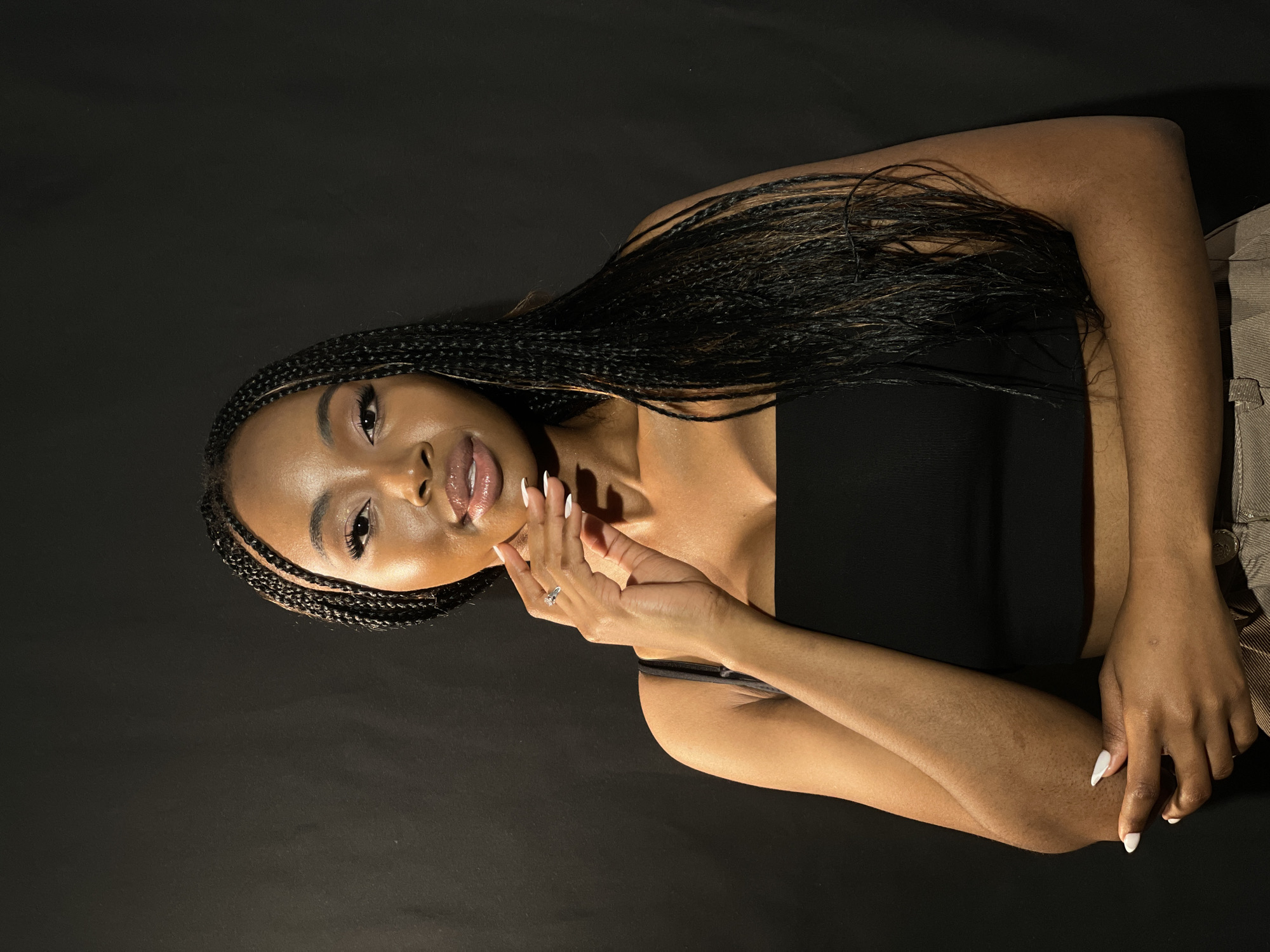 A young Black woman is depicted in a studio portrait, positioned diagonally across a solid black background. She lies or leans back with her head slightly tilted, looking directly towards the viewer with a confident and alluring expression, enhanced by a subtle smile and shimmering lips. Her eyes are open and engaging, framed by defined eyebrows and long eyelashes.
Her hair consists of numerous long, dark micro braids that cascade over her shoulders and down her back. She wears a black strapless top or dress, which contrasts with her warm brown skin tone. Her right hand, adorned with a ring on her ring finger, is gracefully placed near her chin and neck, drawing attention to her long, pointed white nails. A portion of a light-colored, possibly beige or khaki, garment (trousers or skirt) is visible at the bottom right of the frame, suggesting she is wearing a complete outfit.
The lighting is focused and dramatic, highlighting her features and skin, creating some soft shadows that add depth to the image. The plain black background ensures full attention remains on the subject, without any discernible location cues or visible text. The overall atmosphere is one of sophisticated portraiture.