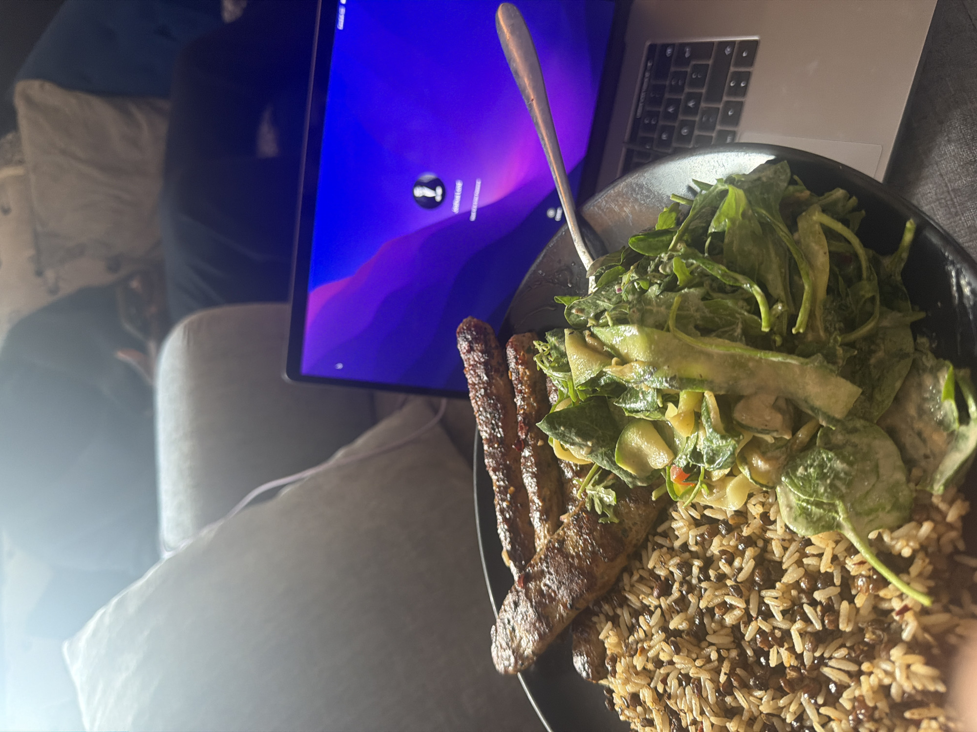 The image shows a bowl of food consisting of rice mixed with lentils, two sausage-like items, and a leafy salad. A silver spoon is in the bowl. The bowl sits next to a closed laptop computer. In the background is a screen displaying the login screen of a device. The scene appears to be set on a grey couch. The location is not discernible from the image.