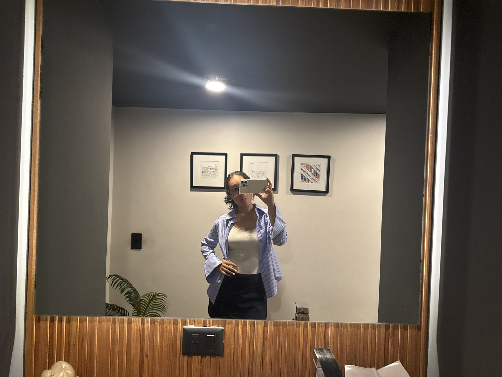 A person stands in front of a mirror, taking a photo of themselves with a smartphone. The person is wearing a white top, a blue and white striped shirt, and a dark skirt. Three framed pictures hang on the wall behind them, and a small plant is visible on the left. The mirror is framed with wood paneling. Below the mirror, there is a light switch and some bathroom supplies. The location is possibly a bathroom or restroom within a building in San Bernardino Tlaxcalancingo, Mexico, as specified.