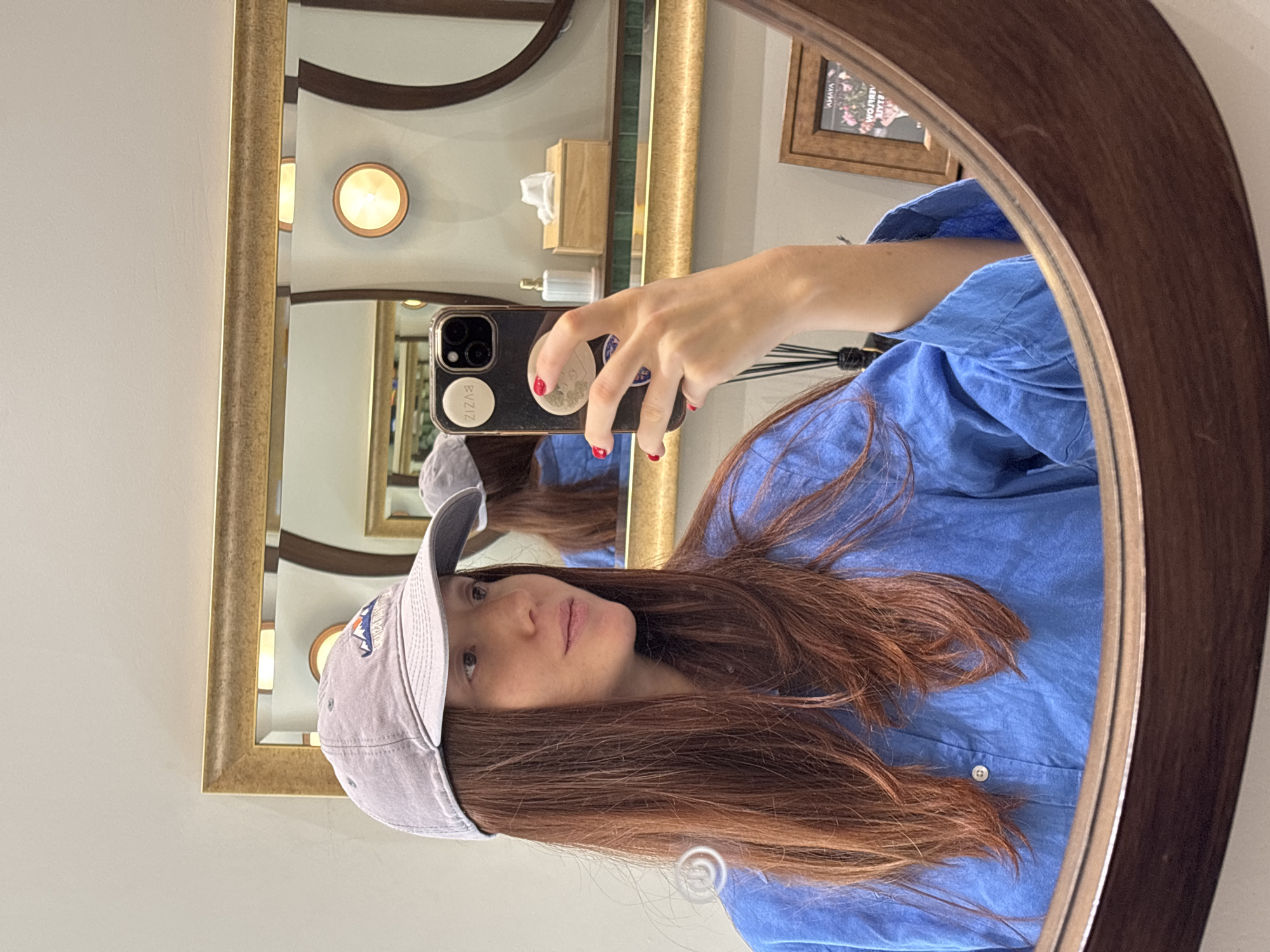A woman is taking a selfie in a mirror. She is wearing a light blue button-down shirt and a gray baseball cap. She is holding a black iPhone with stickers on the back. The mirror has a gold frame and is in a bathroom. There is a box of tissues on the counter. The city is Choeng Thale, Thailand.