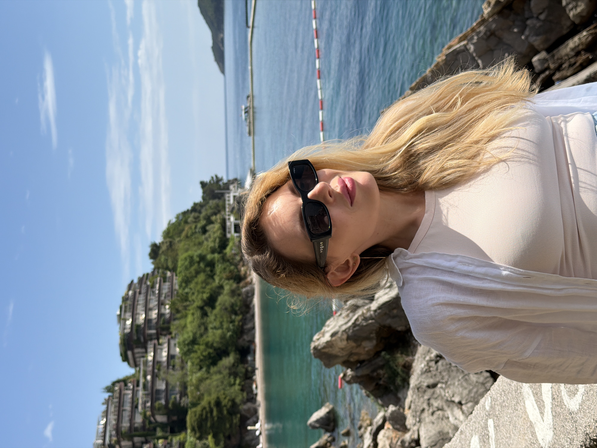 A woman with long blonde hair is shown in a medium shot, wearing dark sunglasses, a light-colored top, and a white blouse. She is standing outside, likely on a rocky ledge overlooking a beach with turquoise water. In the background, there is a row of modern buildings stacked on top of each other, surrounded by lush greenery. The scene appears to be in or near Budva, Montenegro, given the architectural style and coastal environment. The sky is blue with some clouds.