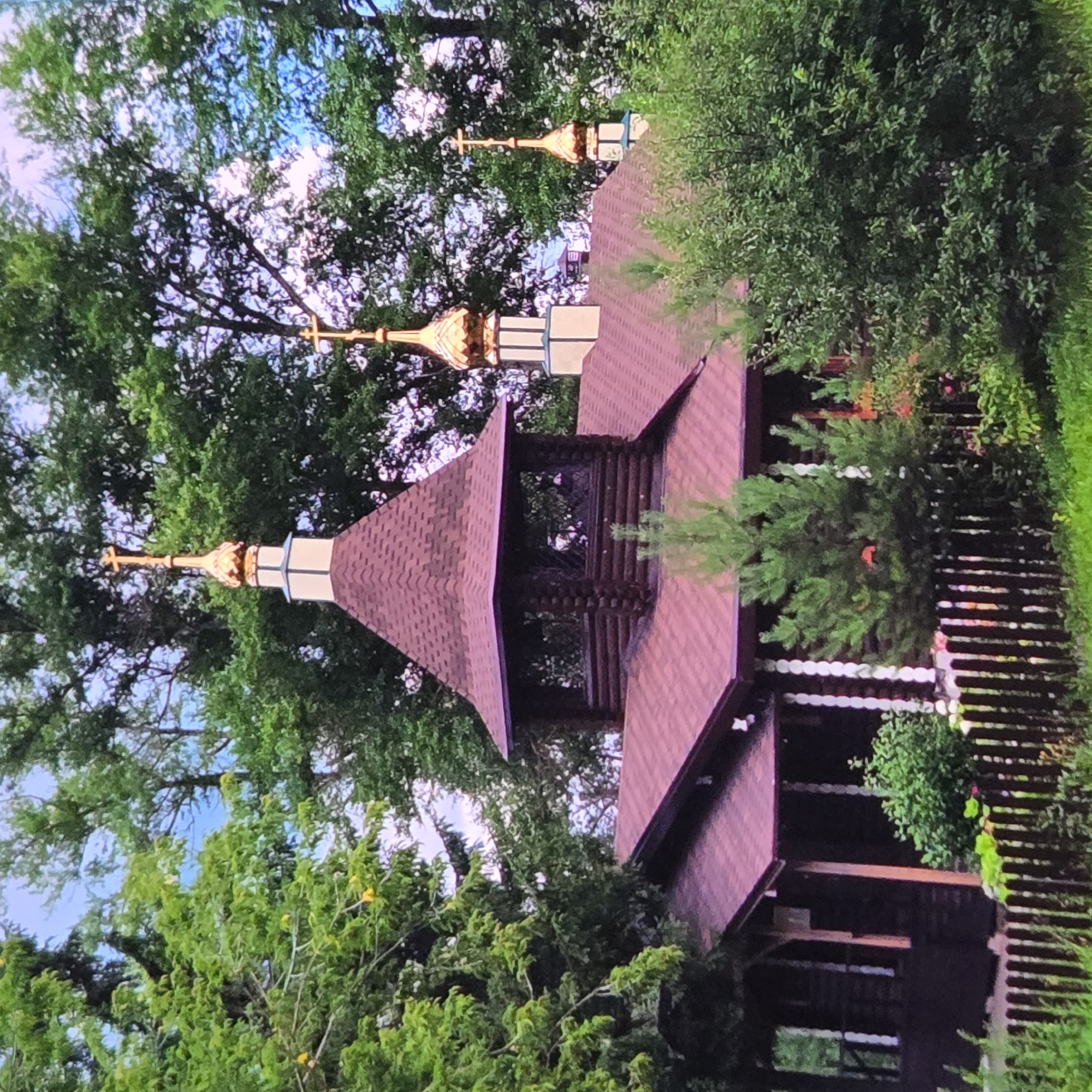 The image shows a small, wooden, Russian Orthodox church nestled amongst lush green trees. The church features a prominent red roof and several golden domes topped with crosses. A wooden fence encloses the property, and dense foliage surrounds the building. The sky is visible through the trees, appearing partly cloudy. The scene suggests a peaceful, rural setting.