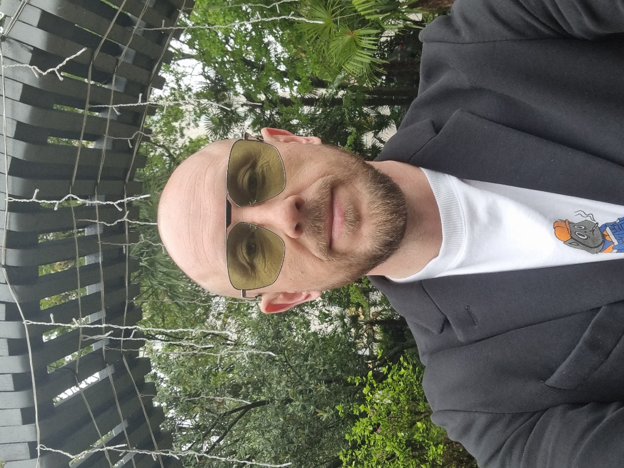 A bald man with a beard and yellow sunglasses is pictured from a low angle, wearing a white t-shirt with a cartoon graphic underneath a black blazer. He is positioned against a background of green trees and foliage, interspersed with what appears to be an arched structure made of dark, horizontal slats, ornamented with strands of small lights. Based on the details of the landscaping and the architectural structure, the location is plausibly within the city of Sochi, Russia.