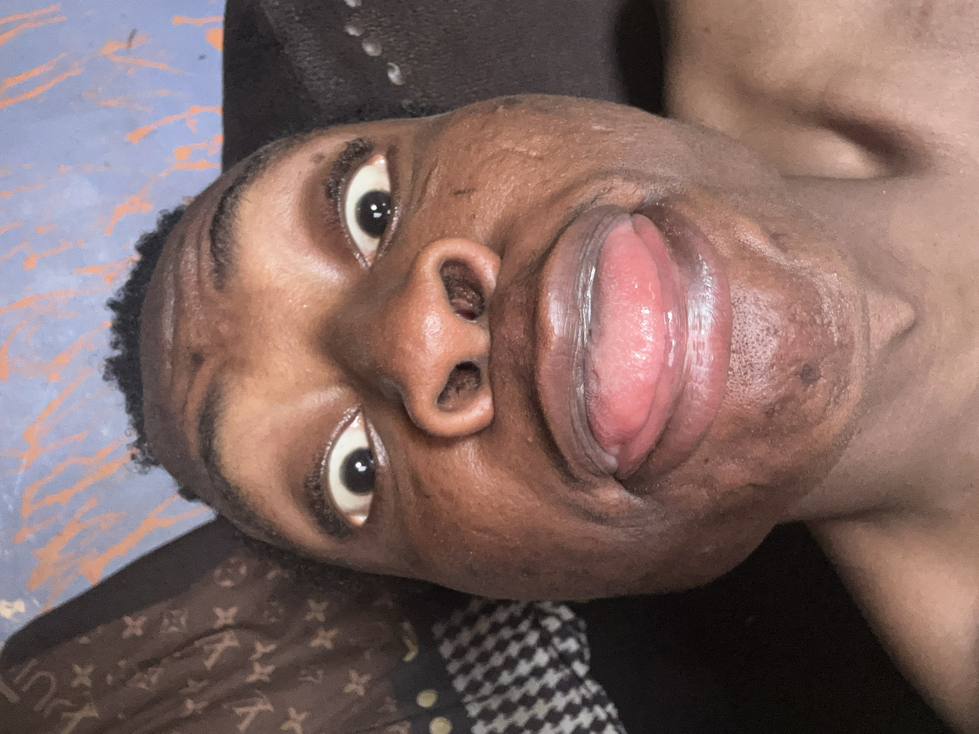The image shows a close-up, low-angle shot of a Black male's face with his tongue sticking out. The subject has short, dark hair and dark eyes. His skin tone is dark brown. Part of his bare shoulder is visible in the upper right corner. The background includes a patterned material near his head and what appears to be a blue and orange painted surface in the upper left corner. The location is not specified.