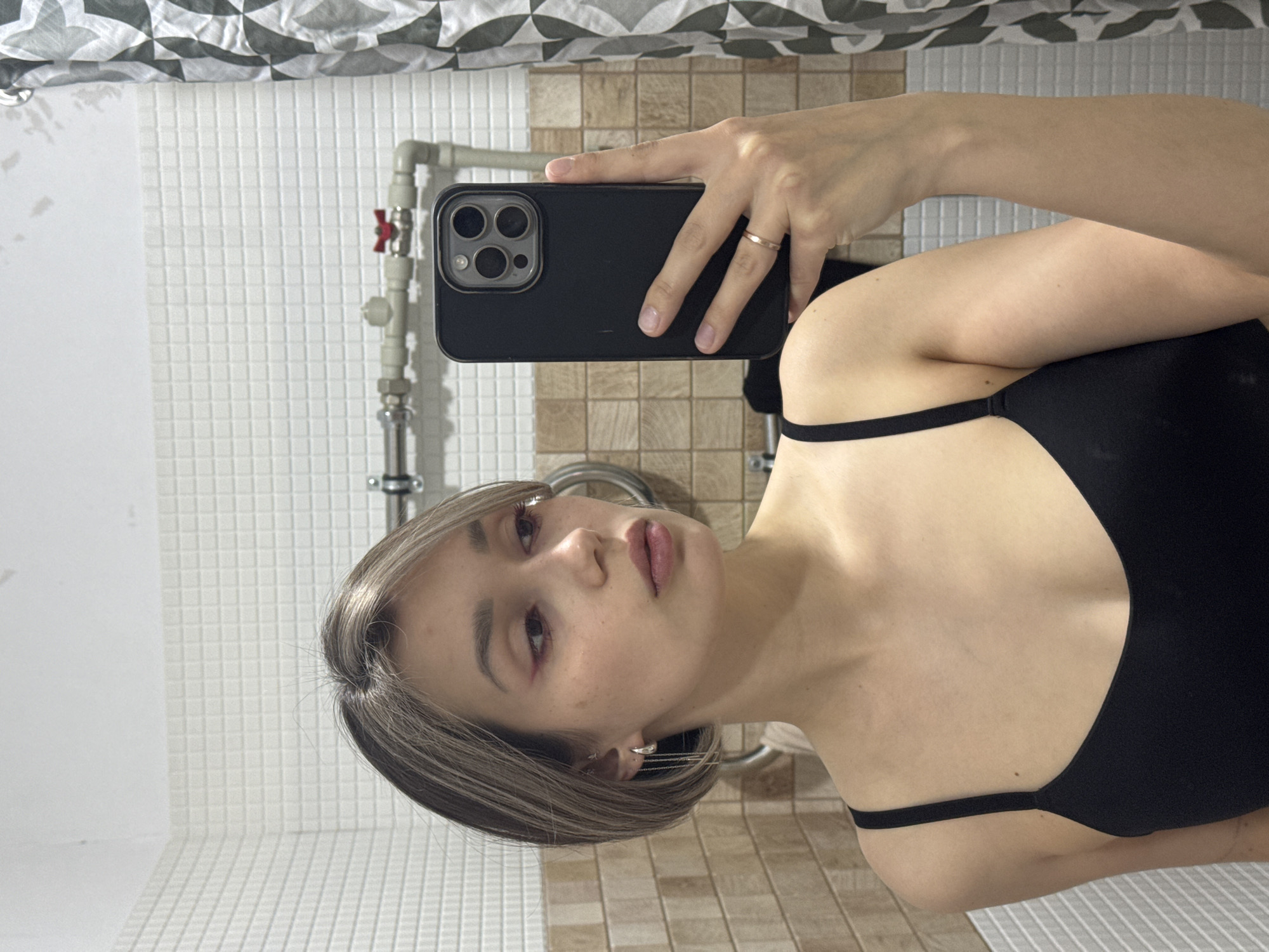 A young woman with shoulder-length blonde hair is taking a selfie in a bathroom. She is wearing a black top. The background includes white tiled walls, beige tiled walls, a shower curtain, and pipes. The location appears to be inside a residential bathroom in Unknown, Unknown.