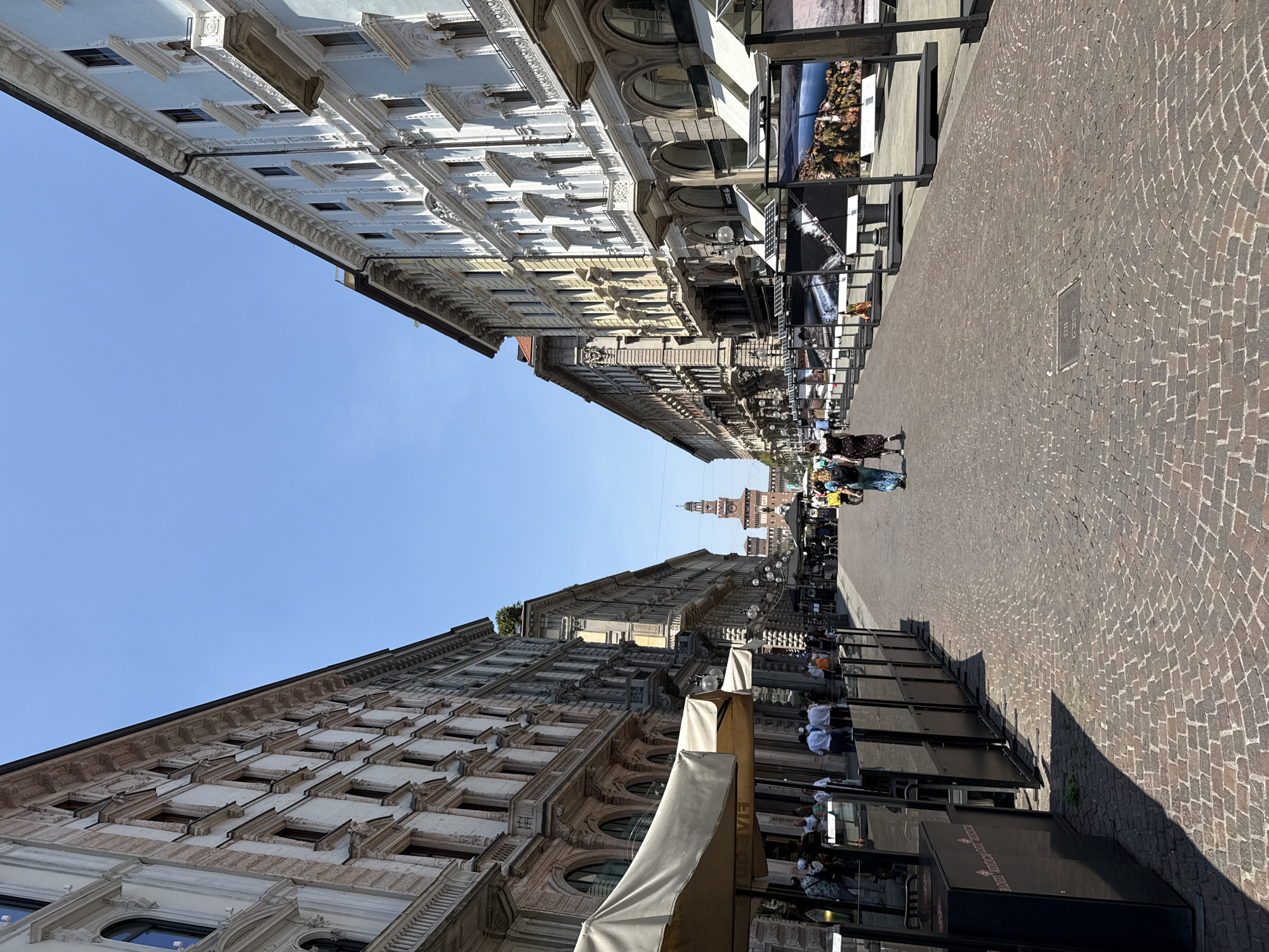 A street scene in a European city on a sunny day, likely Milan, Italy, judging by the Sforza Castle in the distance. The street is paved with cobblestones and lined with ornate buildings on both sides. In the foreground, a cafe with outdoor seating is visible on the left. Further down the street, an exhibition of large framed photographs is displayed on the right. Several people are walking along the street, and some are seated at the cafe. The sky is clear and blue.