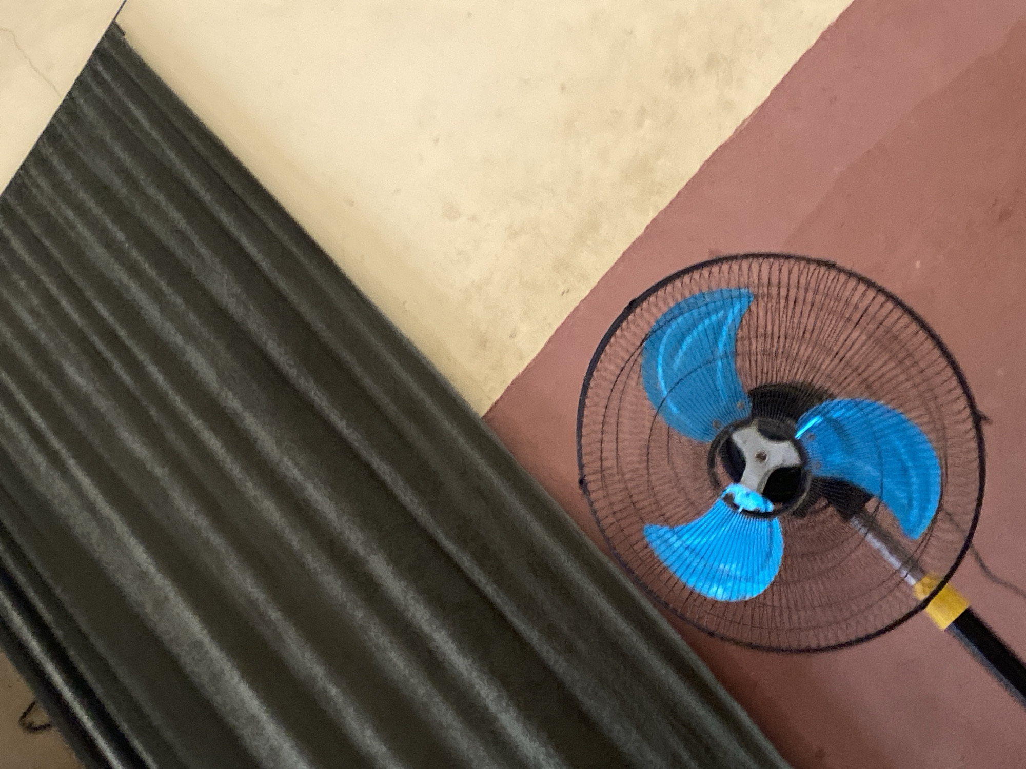 The image shows a close-up, high-angle shot of a black oscillating fan with bright blue fan blades. The fan is positioned in the lower right quadrant of the frame, against a reddish-brown surface that appears to be a floor or wall. The fan's grille is visible, as is the central hub where the blades are attached. A yellow band is visible on the fan's black pole.
The background is divided into two distinct areas. To the left of the fan, a large portion of the image is taken up by dark, ribbed material, likely curtains or a similar fabric, which hang diagonally across the frame. To the top and right of the ribbed material is a light-colored surface, appearing creamy or pale yellow, which has some faint staining or discoloration. The division between the dark ribbed material and the light-colored surface forms a strong diagonal line. The reddish-brown surface upon which the fan sits also forms a diagonal line, meeting the light-colored surface.
The lighting suggests it is daytime, possibly indoors with ambient light. There are no people visible, and the activity is static, simply depicting the fan in its environment. No text is visible in the image. The scene is domestic or possibly a common area in a building in Jauro Yinu, Nigeria.