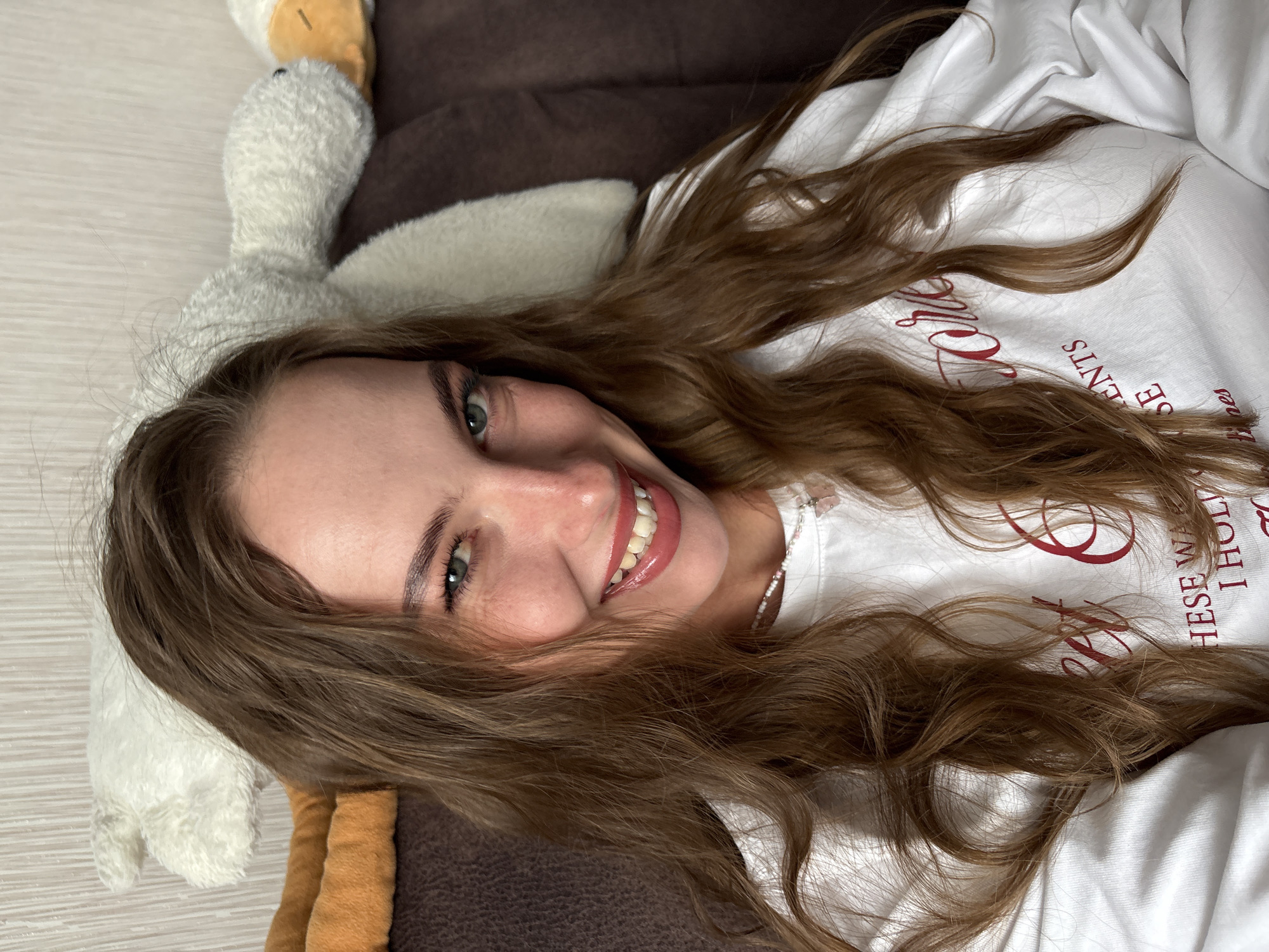 An indoor portrait taken in Perm, Russia, features a young woman with long, wavy, light brown hair and light green eyes, smiling directly at the camera. Her lips are coated with glossy pink lipstick, revealing her teeth, and dimples are visible on her cheeks. She wears a white long-sleeved top with red lettering that partially reads "THESE WAR" and "I HOLD". A white beaded necklace with a small pink pendant is around her neck. The background includes a light-colored, textured wall and a brown upholstered surface where two plush toys are visible: a white, duck-shaped toy with an orange beak and an additional orange/brown plush object.