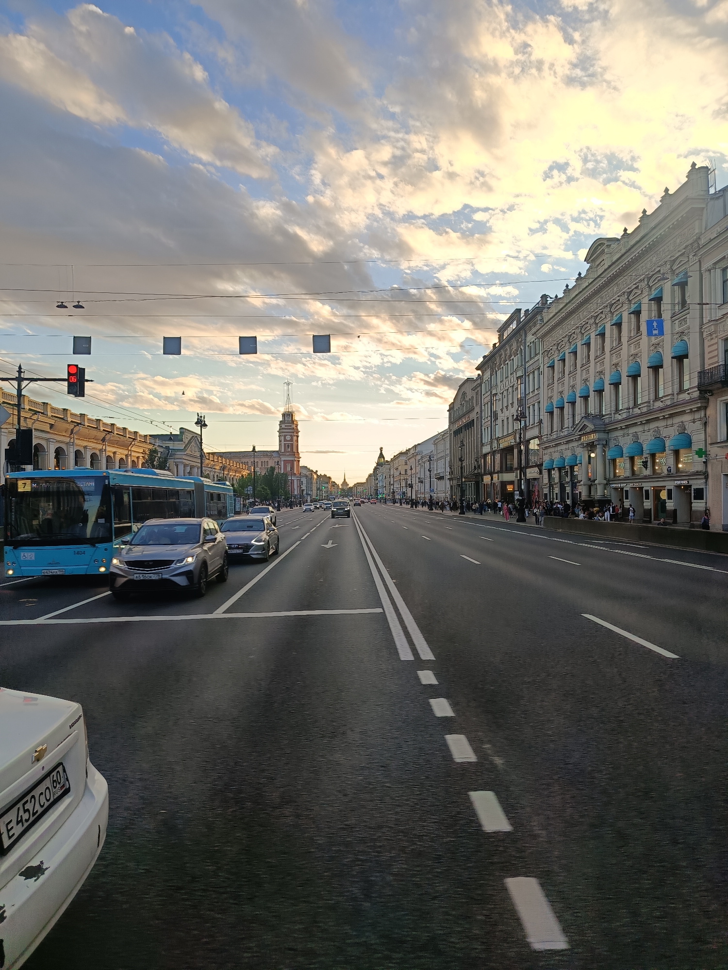 The image depicts a wide, multi-lane city street with traffic. On the right side of the street, a row of ornate, multi-story buildings with blue awnings lines the sidewalk. Pedestrians are visible on the sidewalk in front of these buildings. On the left side of the street, a blue bus is stopped at a traffic light, which is red. Several cars are in motion and parked. Overhead, power lines and square boxes are suspended across the street. The sky is filled with dramatic, illuminated clouds. In the distance, a tall clock tower with a spire is visible. The scene appears to be captured at dusk or during golden hour.