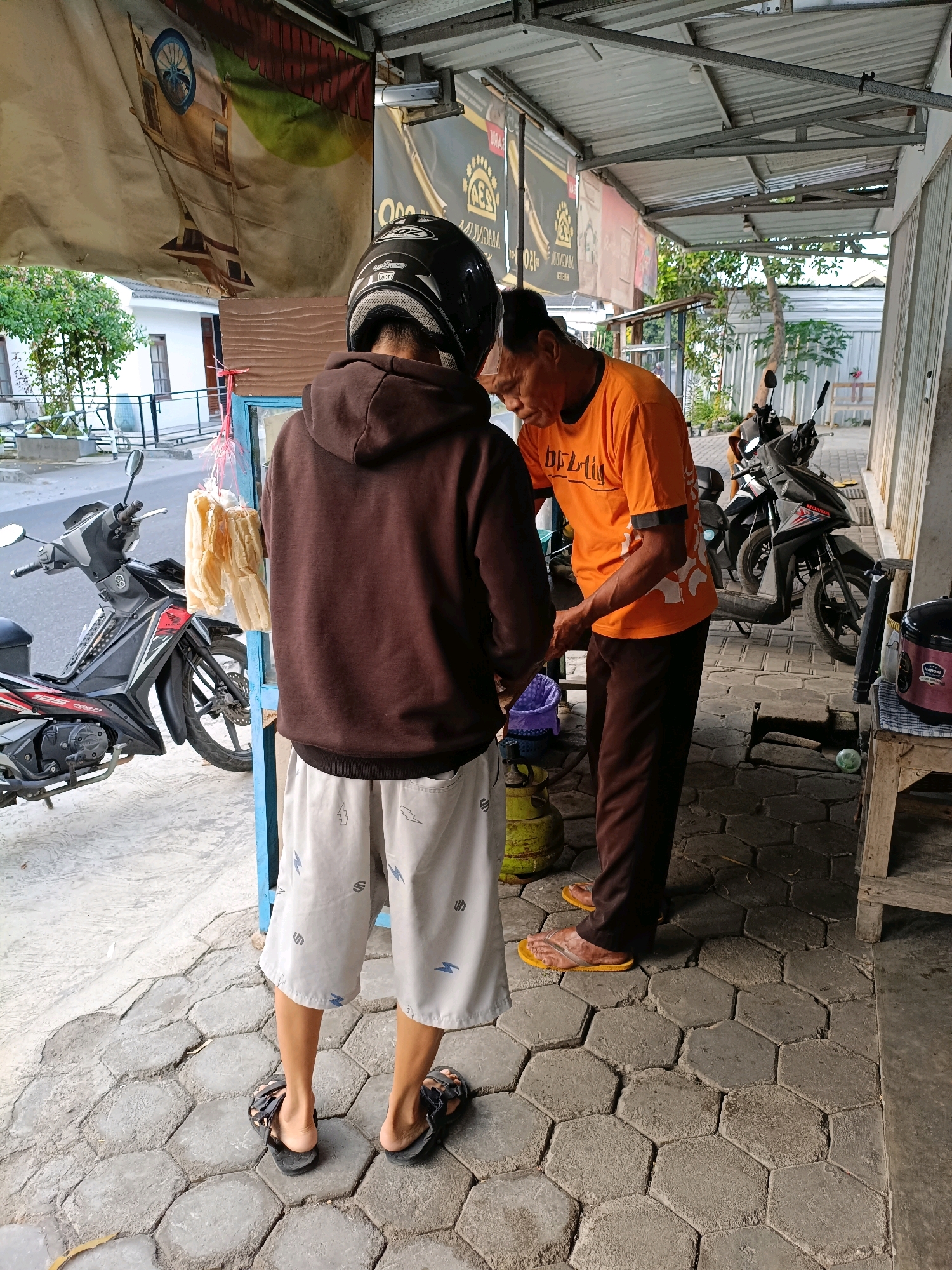 The image depicts a street scene in Maguwoharjo, Indonesia, focusing on two individuals near a small outdoor stall. In the foreground, a person wearing a brown hooded jacket, white shorts with a lightning bolt pattern, and black sandals stands with their back to the camera, facing a man in an orange t-shirt and brown pants. The man in orange appears to be interacting with something in his hands, possibly preparing food or handling merchandise. He is wearing yellow flip-flops. To the left of the scene, a black motorcycle is parked. The ground is paved with hexagonal concrete blocks. The stall itself is partially visible, with a metal roof supported by a structure. Behind the stall, more motorcycles are parked, and vegetation is visible. A banner with a boat graphic hangs above the stall area. A yellow gas cylinder is also present on the ground near the stall. The overall environment suggests a bustling commercial area within a residential or mixed-use part of the city.