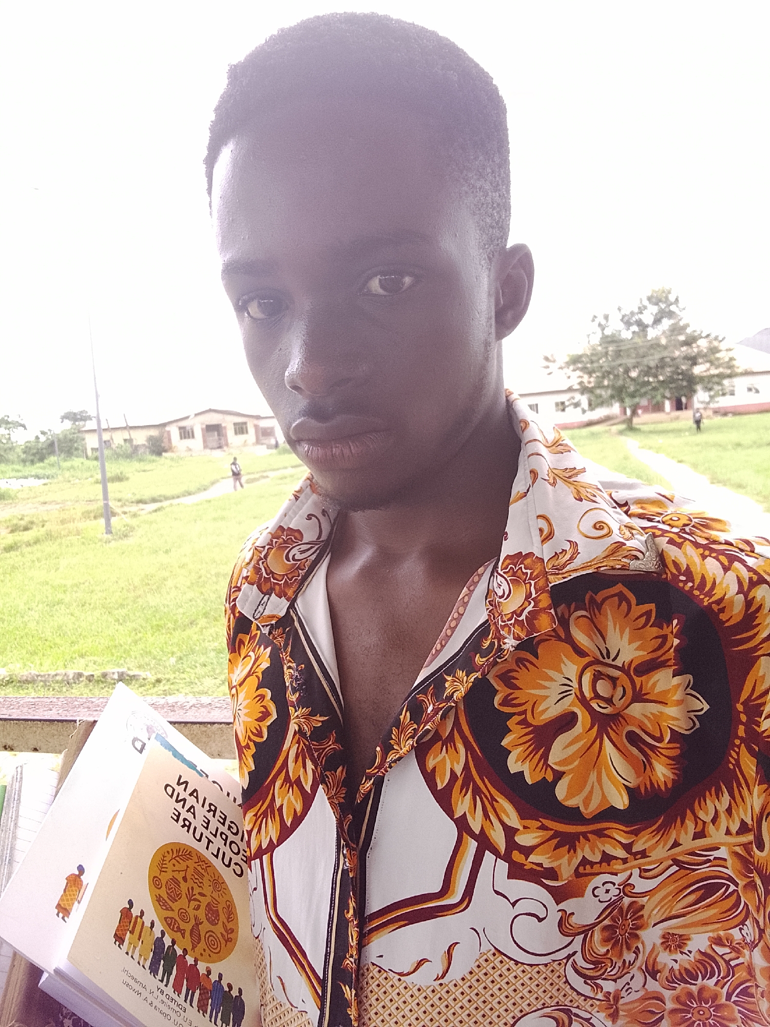 A young Black male is taking a selfie outdoors. He is wearing a collared, short-sleeved shirt with a busy, ornamental pattern of orange, yellow, and brown floral designs on a white background. In his left hand, he holds open a book titled "GERMAN PEOPLE AND CULTURE." The background shows a slightly overcast sky above a grassy field with a few buildings and trees. In the middle ground, a path winds through the grass, and a person can be seen walking on it. In the distance, there are more buildings and vegetation, consistent with the landscape of Ihiagwa, Nigeria. A railing or ledge is visible in the foreground.