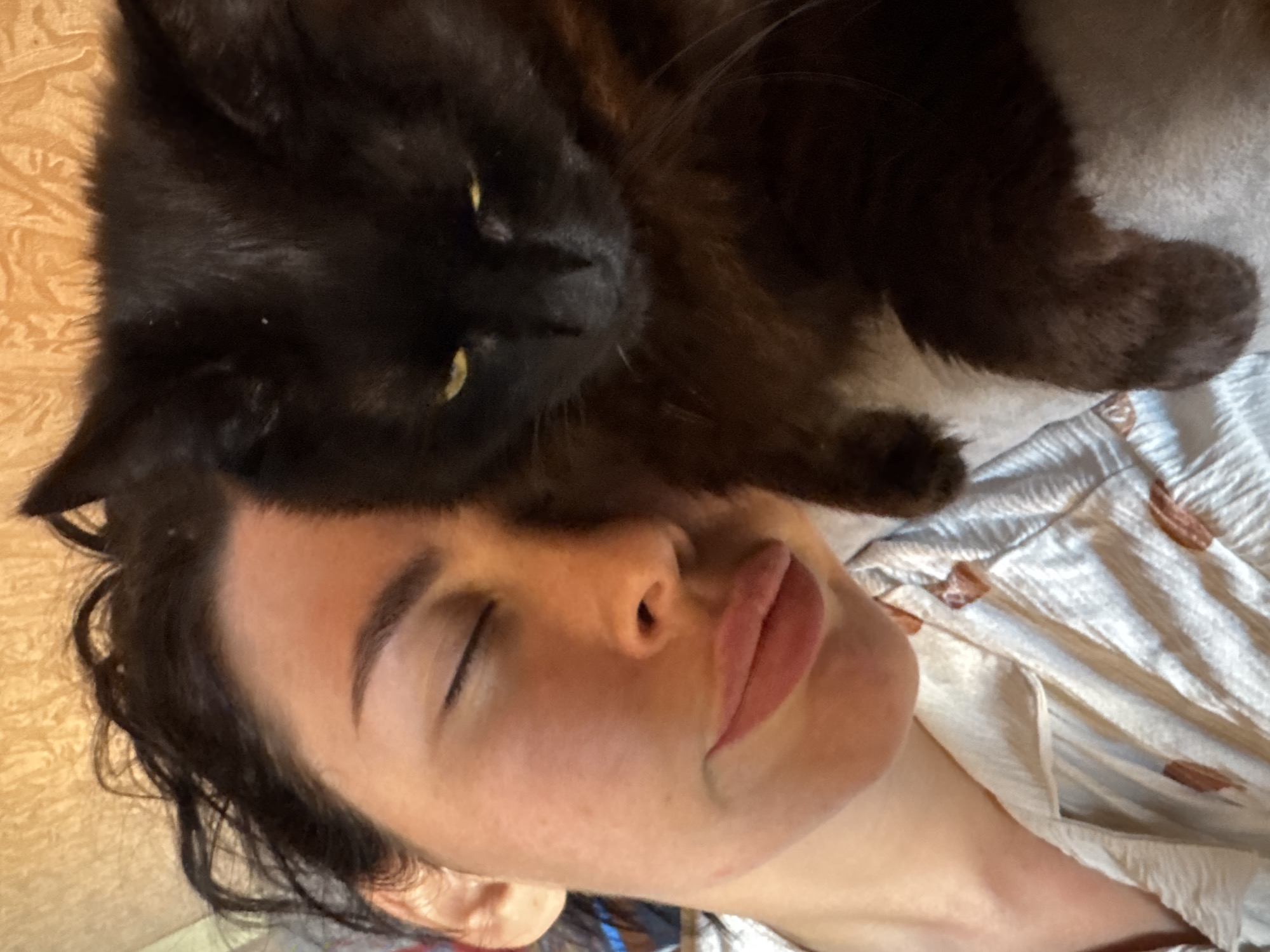 A woman is lying down with her eyes closed. A black cat is lying on her head. The woman is wearing a patterned shirt. The background shows a wooden surface. The location is not discernible from the image.