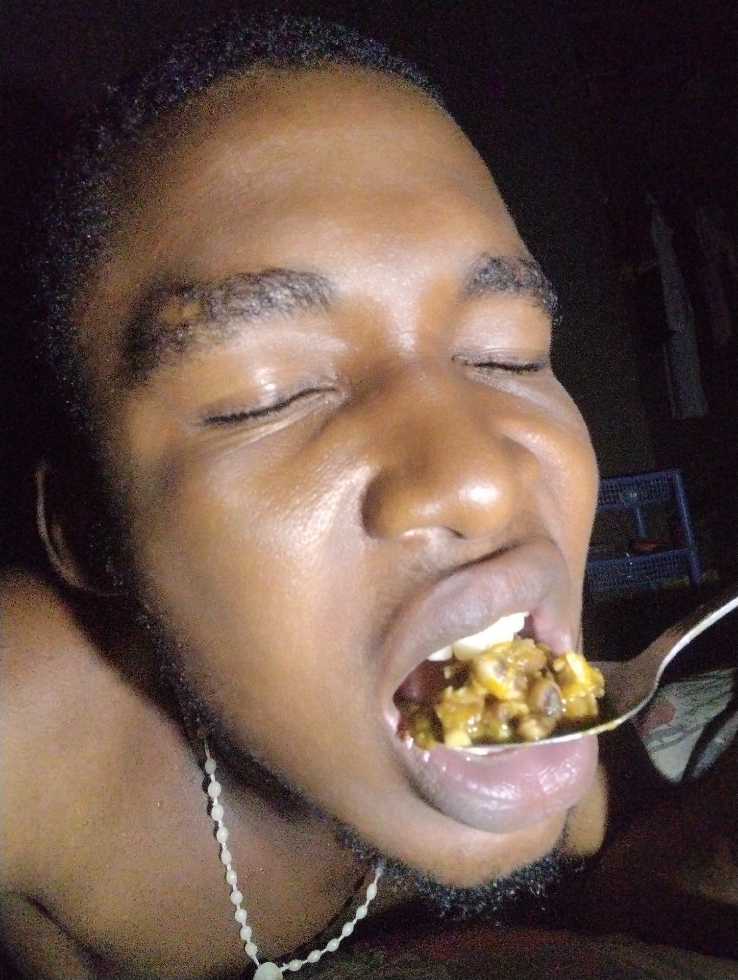 The image shows a close-up, low-angle shot of a person with their eyes closed, about to eat from a spoon. The person has dark skin and short, dark hair. They are wearing a white beaded necklace. The spoon contains a portion of food that appears to be a mixture of small, yellowish-brown pieces. The background is dark and out of focus, but a blue plastic shelf unit is visible on the right side. The image appears to have been taken indoors, likely in a residential setting in Ihiagwa, Nigeria.