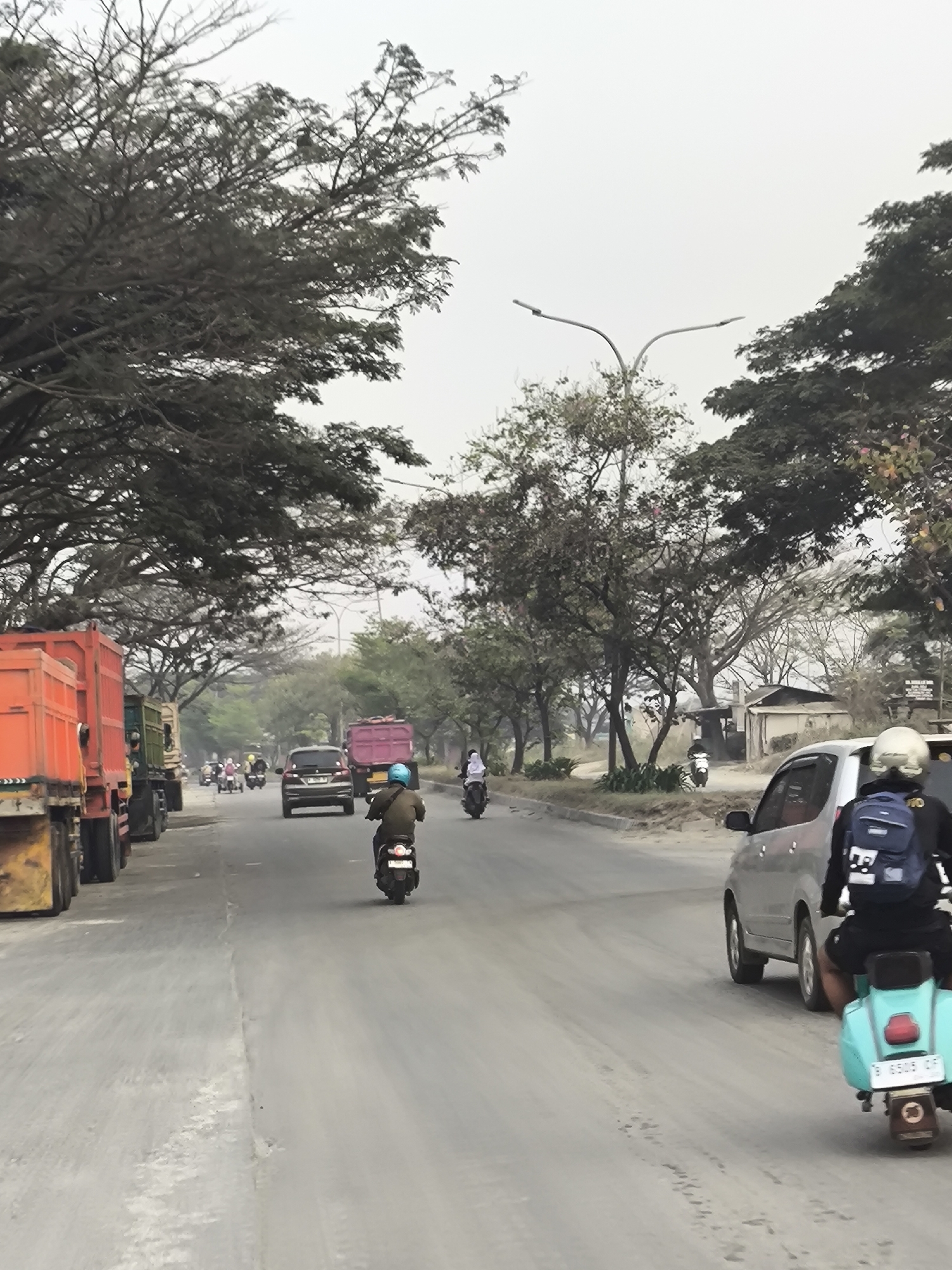 The image depicts a street scene in Cilegon, Indonesia, characterized by overcast skies and a hazy atmosphere. The road is paved and appears dusty, with several vehicles present. On the left side of the road, a row of large orange and red trucks are parked or moving slowly. Further down the road, a pink truck is also visible. Several motorcycles and a car are on the road, with some individuals riding them. One person on a motorcycle wears a blue backpack. On the right side of the road, a silver car is visible, and behind it, a turquoise scooter with a rider wearing a helmet and a backpack. Trees line both sides of the road, with some bare branches and others with dense foliage, suggesting a dry season or a particular type of vegetation. In the background, there are some low-lying buildings and more trees. The overall impression is of a somewhat industrial or commercial area with moderate traffic.