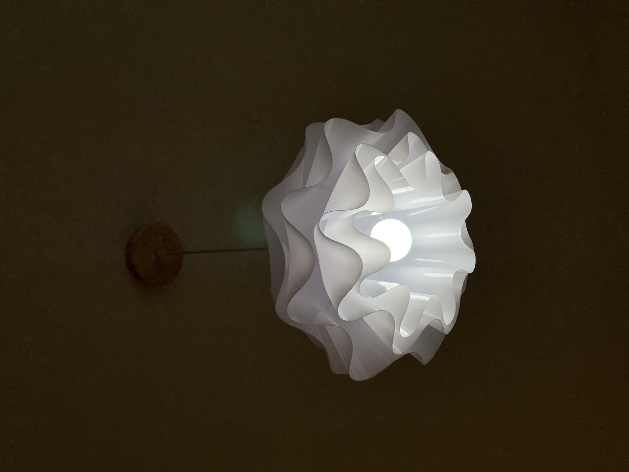 A low-angle shot depicts a white pendant lamp illuminating a room with dark brown walls. The lamp features a spherical bulb enveloped by layers of undulating white material, creating a soft, diffused glow. A cord extends from the lamp to a circular mounting fixture on the wall. The scene takes place in a city called Unknown, Unknown.