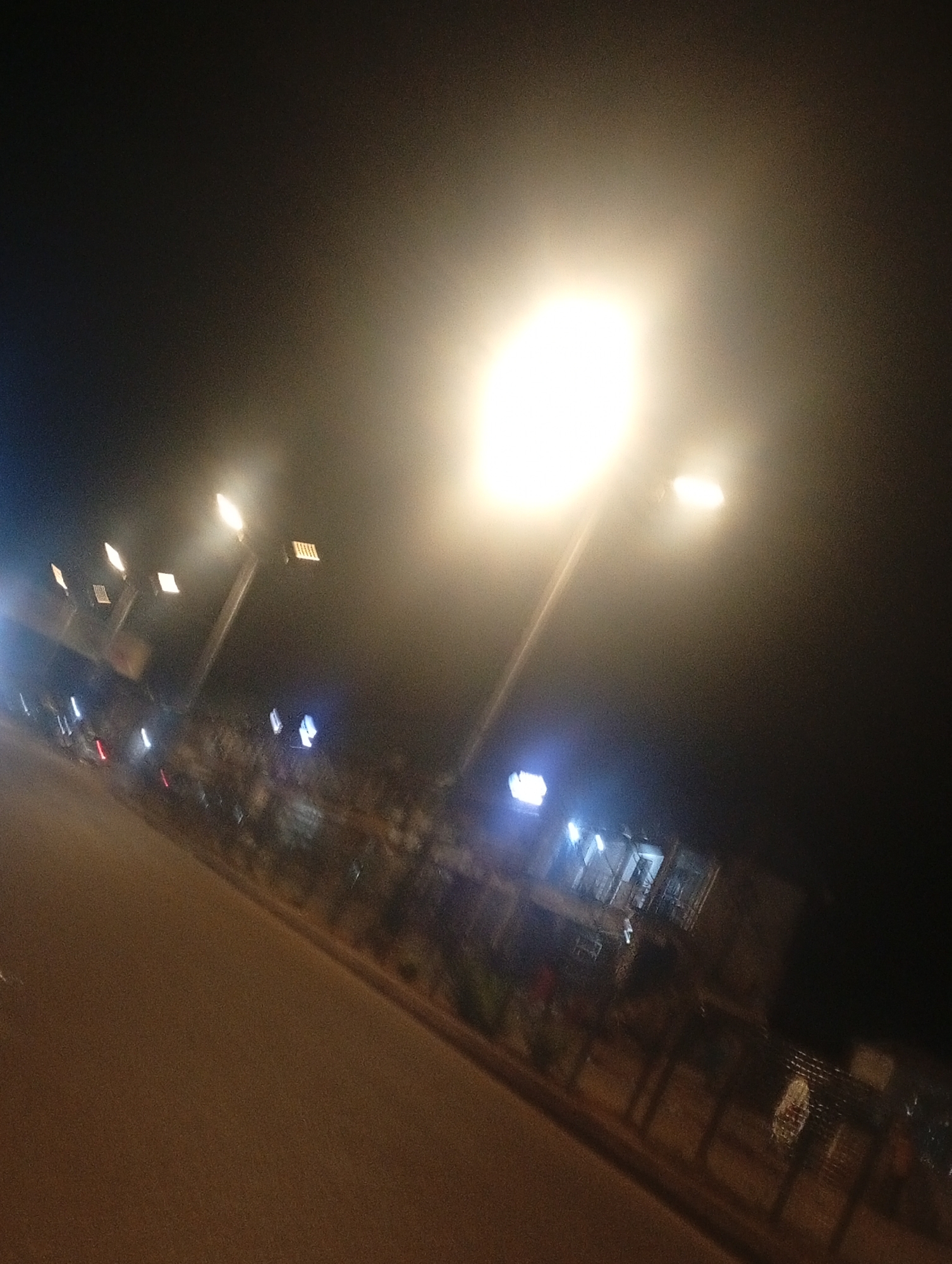 The image depicts a street scene at night in Bukuru, Nigeria, illuminated by streetlights. A paved road dominates the foreground, curving slightly to the right. Along the right side of the road, there is a sidewalk or pedestrian area with some vegetation and what appear to be buildings or structures. Numerous streetlights are visible, casting a bright glow and creating a hazy effect due to the night and possibly atmospheric conditions. Some of the lights are mounted on poles, while others appear to be integrated into structures. There are indistinct shapes that could be vehicles or people on the road or in the background, but they are not clearly defined. The overall impression is of an urban environment at night, with the streetlights providing the primary source of illumination.