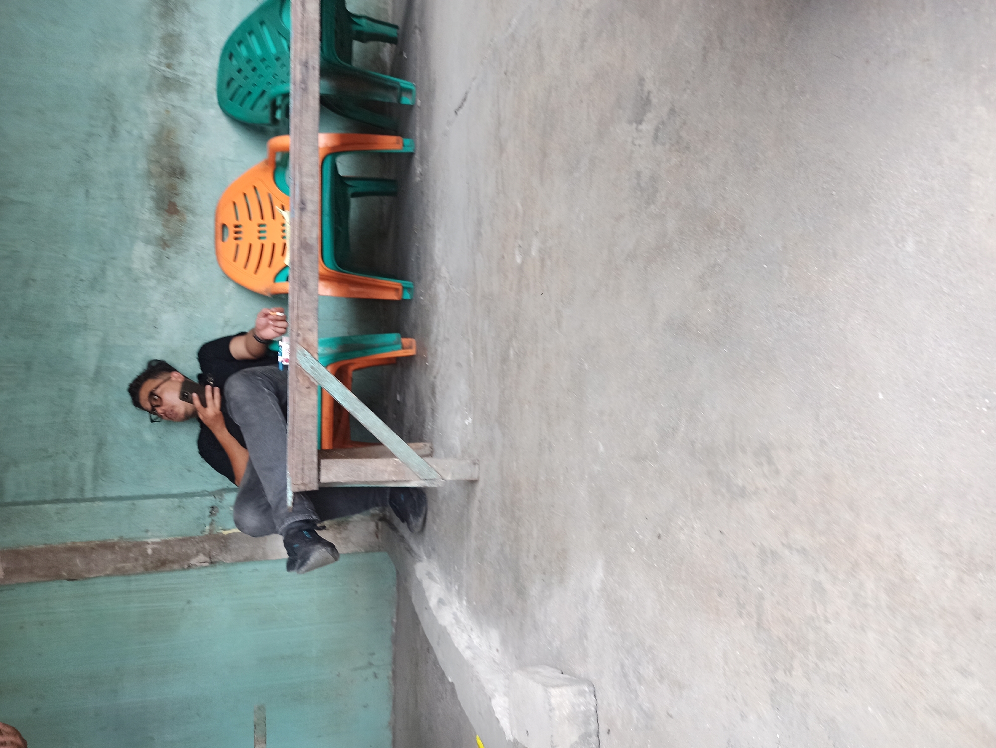A man with dark hair and glasses is sitting on a wooden bench, leaning back and holding a smartphone in his right hand. He is wearing a black t-shirt and dark grey pants. His left leg is crossed over his right, and he is wearing black shoes. In front of him, there are three plastic chairs: two green and one orange. The chairs are placed under a wooden table, which is partially obscured by the man's legs and a diagonal wooden support. The background consists of a teal-colored wall. The ground is made of concrete. The scene appears to be outdoors, possibly in a public area or a small establishment in the city of Medan, Indonesia.