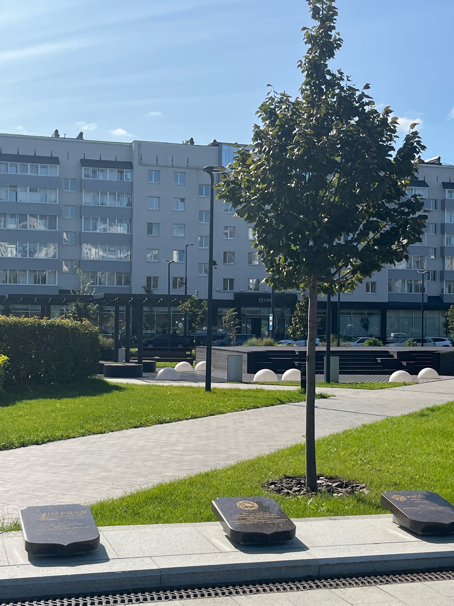 The image shows a contemporary city park area in Perm, Russia, with a modern apartment building in the background. In the foreground, there are three dark, rectangular plaques placed on a light-colored concrete base. These plaques appear to be commemorative, possibly memorial stones, with inscriptions and emblems on them. The plaque on the left has a shield-like emblem at the top and Cyrillic text below. The middle plaque also has a circular emblem with Cyrillic text. The rightmost plaque features a circular emblem with what looks like a globe or a stylized sun and the word "PUCCI" in Latin script.
The park area features well-maintained green grass, paved walkways, and a young tree planted in the center foreground, its shadow falling on the plaques. In the midground, there's a covered seating area with a dark roof and pillars, flanked by dark circular planters. Several white, semi-spherical decorative elements are positioned between the plaques and the covered structure. Streetlights and security cameras are visible along the walkway. The apartment building in the background is multi-story, with many windows and balconies, painted in neutral colors, suggesting a residential area. The sky is clear and blue with a few wispy clouds, indicating fair weather. The scene is calm and orderly, indicative of a planned urban public space.