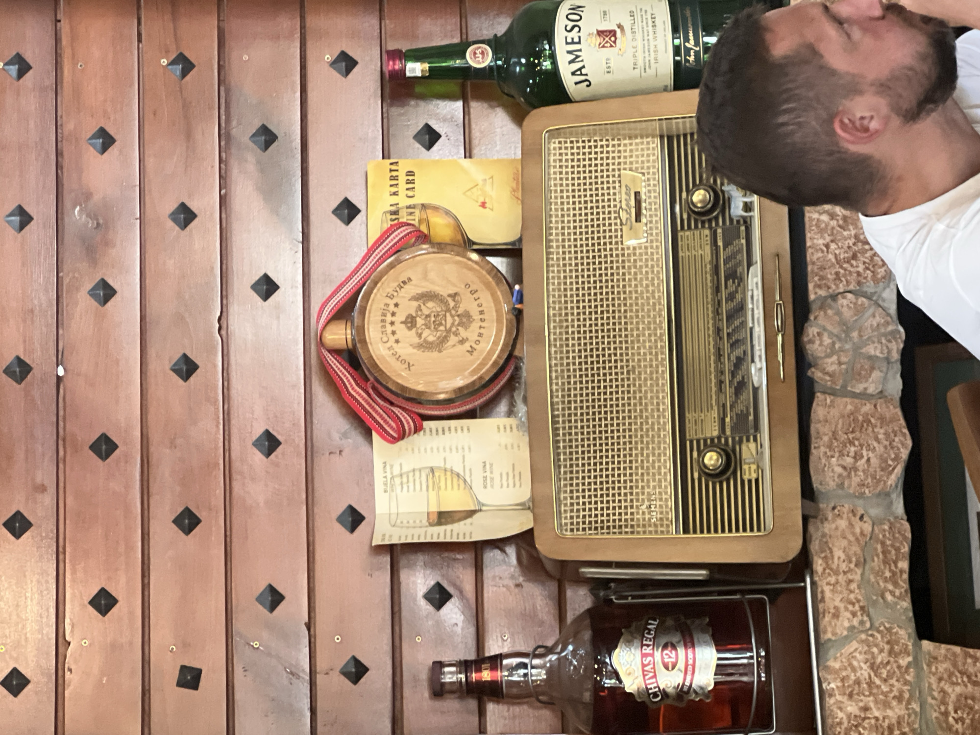 This is a medium-shot, eye-level, indoor photograph of a display shelf in Budva, Montenegro, featuring a vintage radio, two bottles of whiskey, and decorative items. The shelf is constructed from dark wooden planks with a diamond pattern of dark metal studs.
Prominently displayed is a retro-style radio with a gold grille and wooden casing. To the left of the radio sits a bottle of Chivas Regal 12 Year Old Scotch Whisky in a clear glass bottle with a red and gold label. To the right of the radio is a green glass bottle of Jameson Irish Whiskey with its distinctive label.
Above the radio, a round wooden flask or canteen is suspended by a red and white striped ribbon. The flask has an emblem with Cyrillic text and stars, and appears to be from Montenegro. Next to the flask, a yellow card with Cyrillic text, partially obscured, is visible. To the left of the flask, a paper menu or wine list with an illustration of a glass of white wine is propped up.
The shelf is built upon a rustic stone structure, possibly a fireplace or decorative wall. In the foreground, the shoulder and head of a man with short brown hair and a beard are visible on the right side of the frame. He is wearing a white t-shirt and appears to be looking to his right, out of frame. The lighting suggests an indoor setting with ambient light.