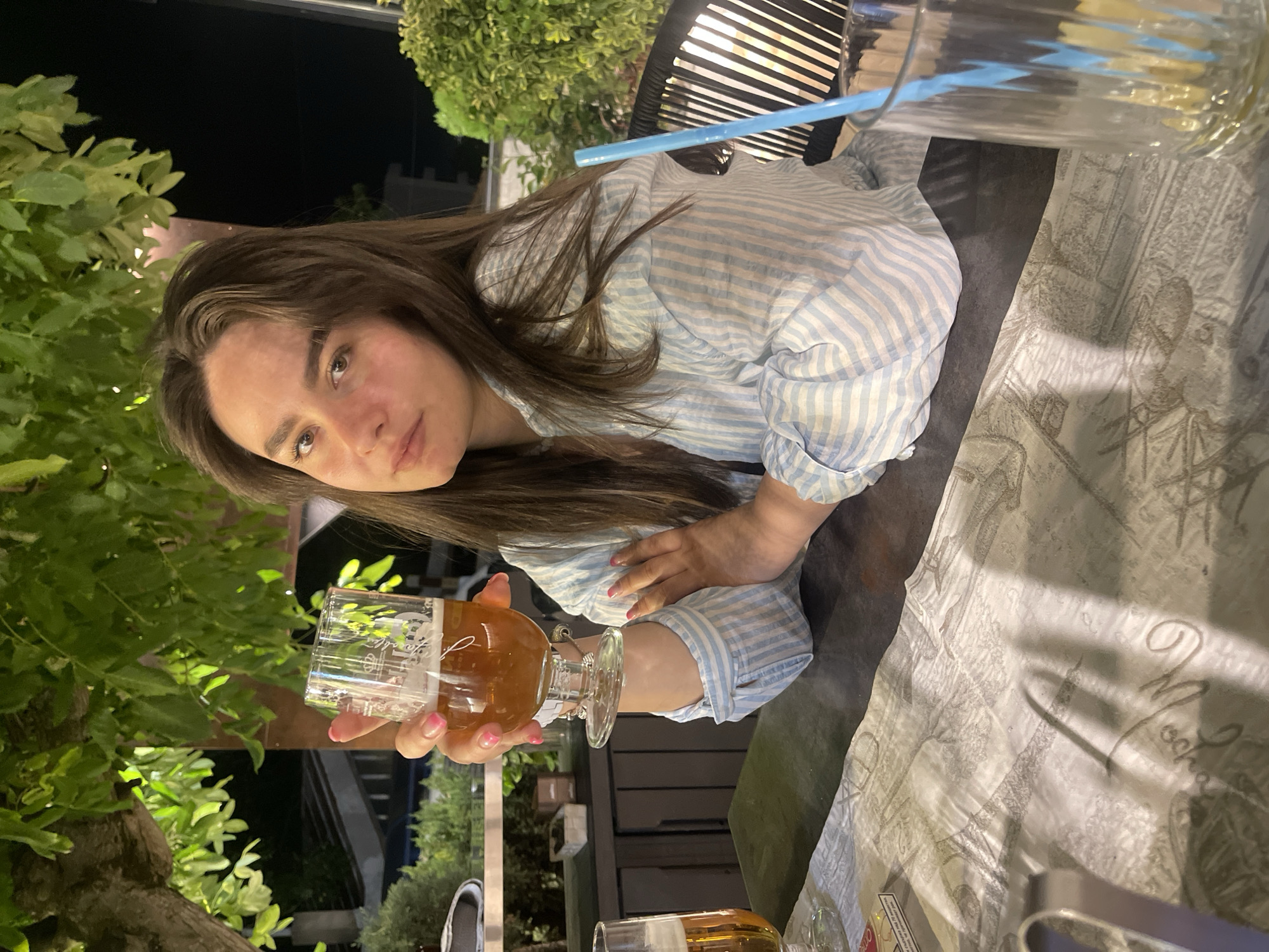 A young woman with long, brown hair and light skin is seated at an outdoor table, holding a glass of beer. She is wearing a light blue and white striped shirt. The table is covered with a patterned cloth. In the background, there are trees, plants, and what appears to be a building. The lighting suggests it is evening. The scene is likely in Sandanski, Bulgaria, as indicated by a clue.